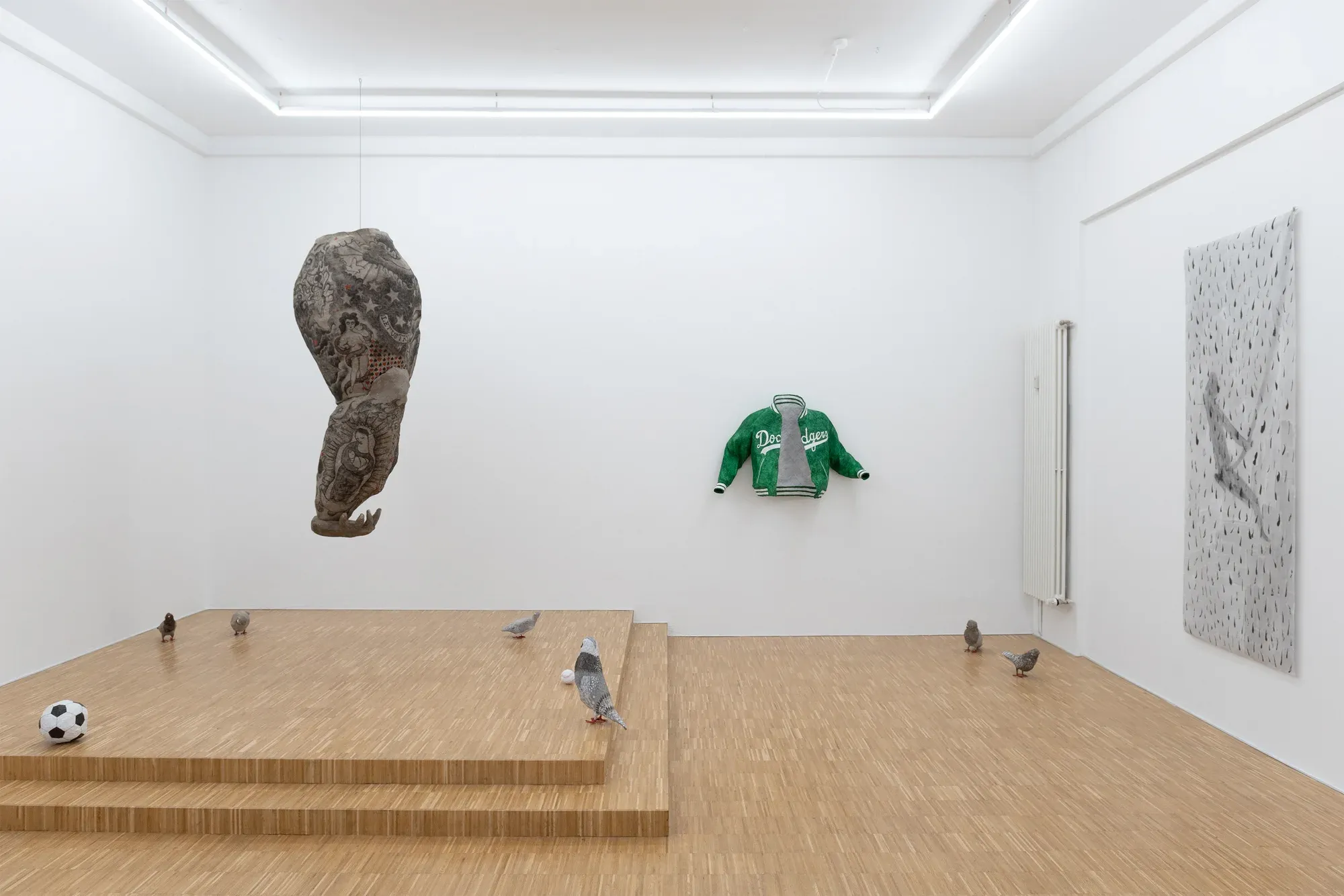 Installation view of a gallery space with a suspended sculptural form, a wall-mounted jacket, floor-based pigeon sculptures and a raised wooden platform.