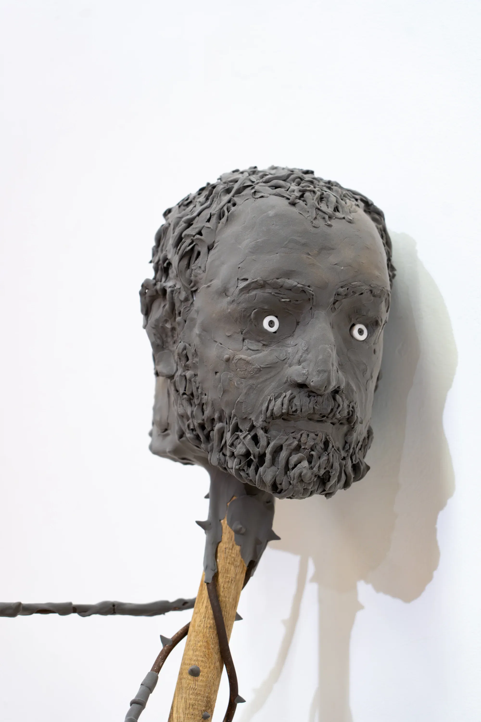 Yorgos Agrotes sculpture The Scarecrow (2025), grey sculpted head mounted on wood and metal structure with visible wires and white eyes.