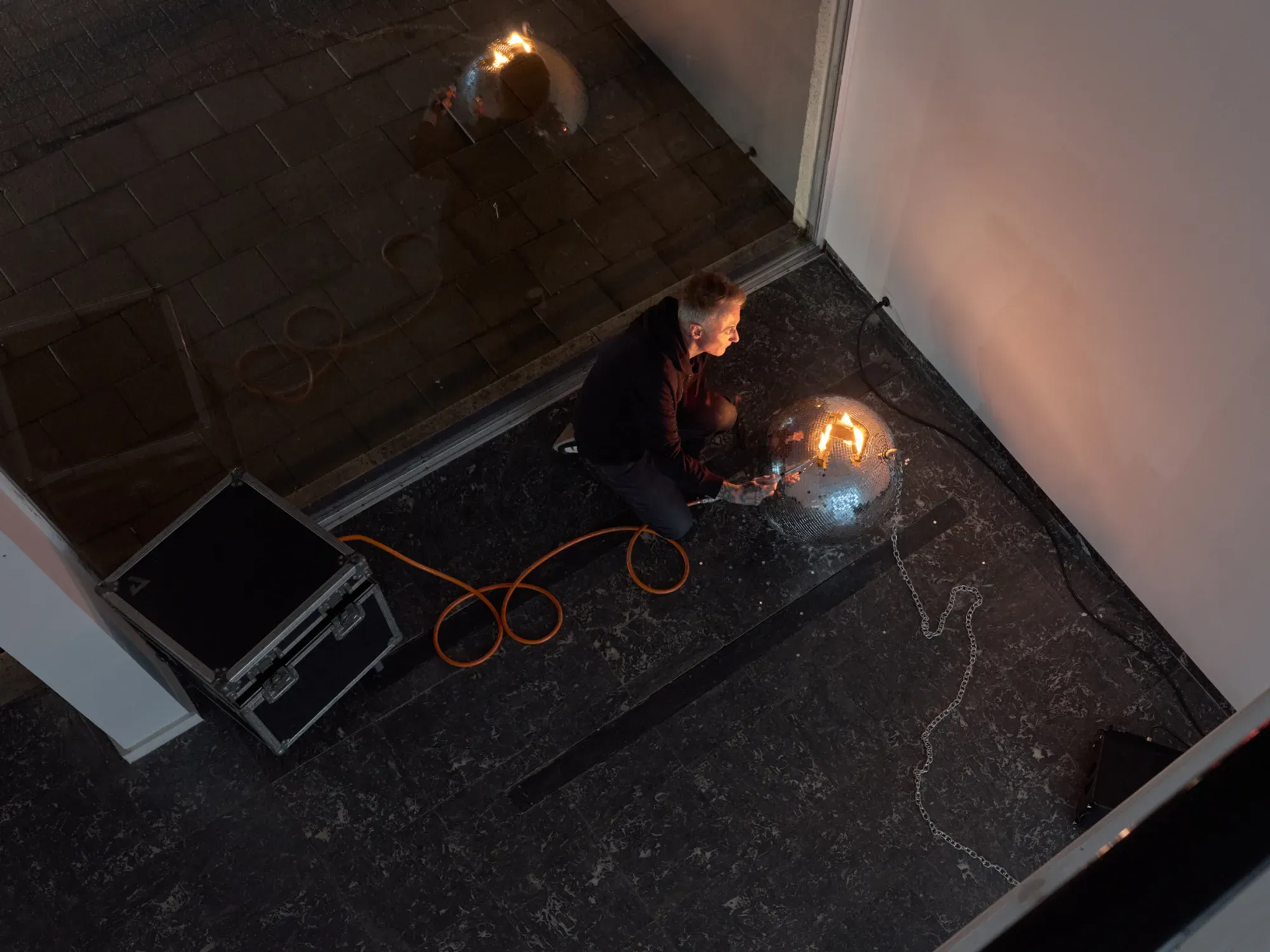 Banks Violette installing and lighting Untitled (Discoball) during the build-up of Wish You Were Here at TICK TACK, Antwerp. The artist kneels beside a mirrored sphere ignited with flame inside the gallery space.