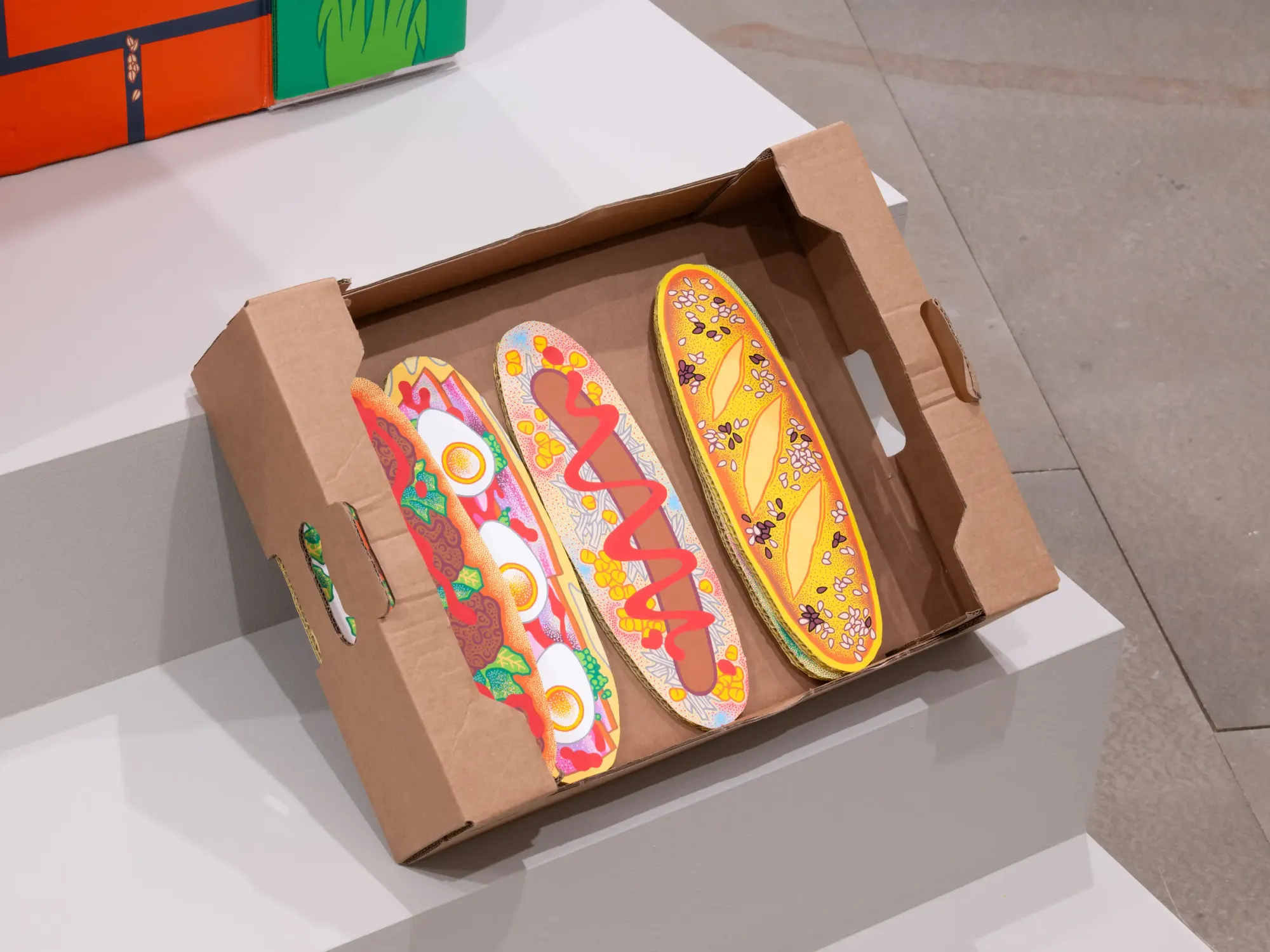 Cardboard box with illustrated baguettes and stylized food imagery, bright colors and hand-drawn stippling detail, Daniela Ponomarevová sculpture