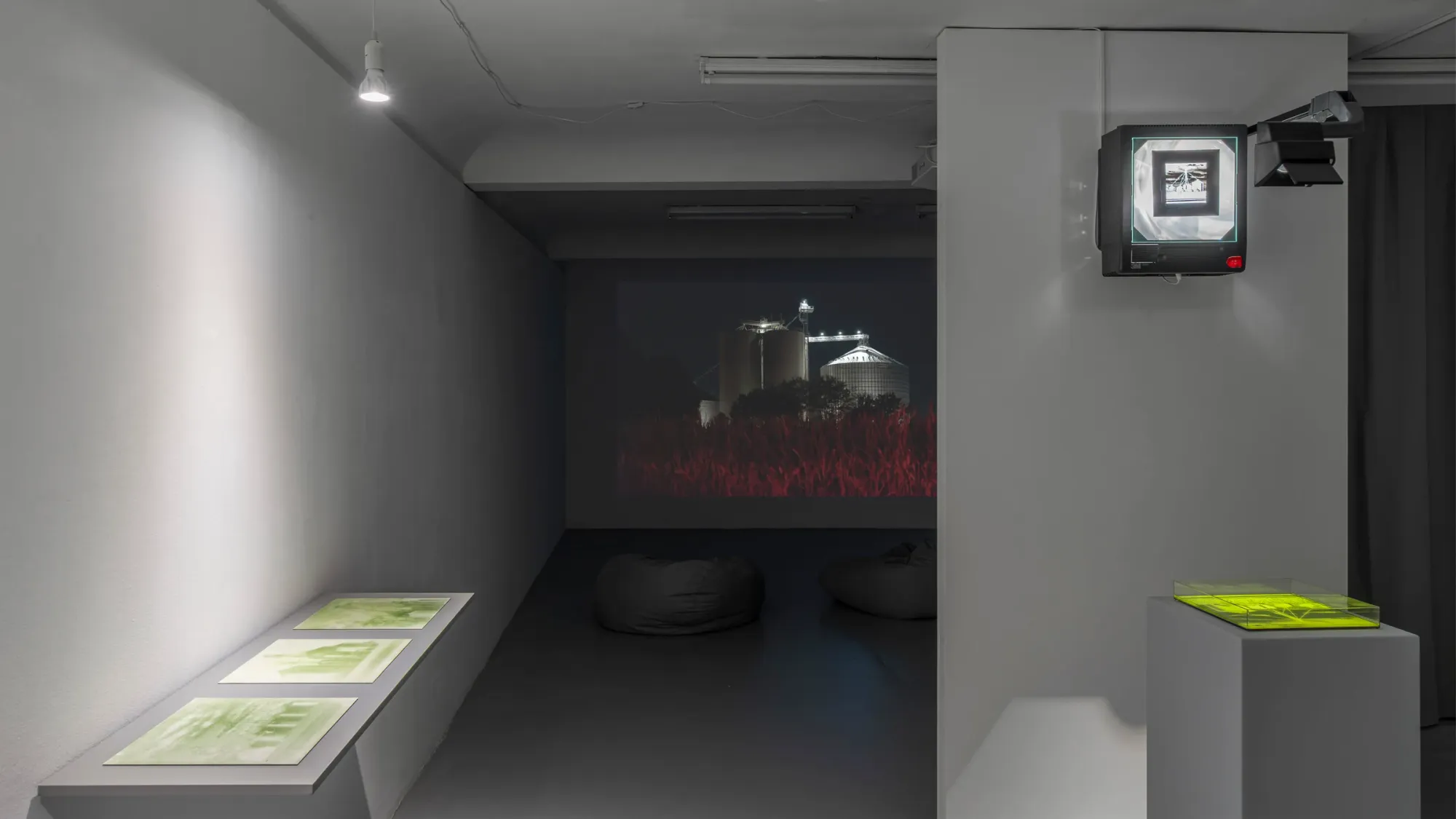 Darkened viewing space with projection of industrial structures in background, beanbags for seating, foreground shows green translucent object and CRT monitor, Kaya & Blank Bloom basis Frankfurt 2026 installation view