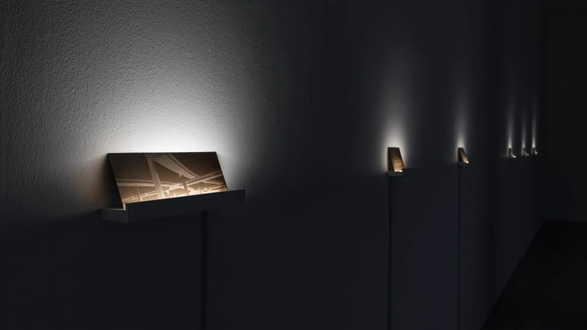 Series of small illuminated photographic works mounted on narrow stands along dark gallery wall, warm light highlighting industrial structures and creating intimate viewing sequence, Kaya & Blank Crude Aesthetics basis Frankfurt 2026 detail