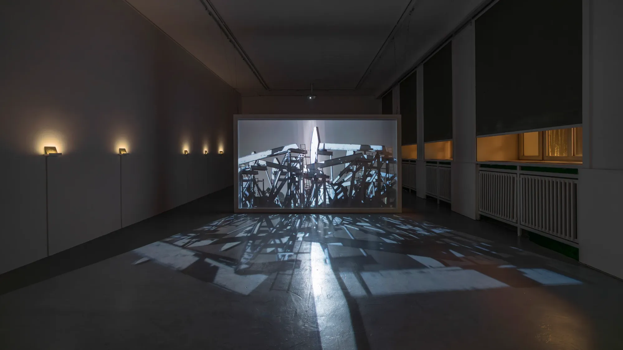 Large illuminated video installation showing multiple oil pump jacks in dark gallery, strong projected shadows spreading across reflective floor, row of small lit works along wall, Kaya & Blank Crude Aesthetics basis Frankfurt 2026 exhibition view