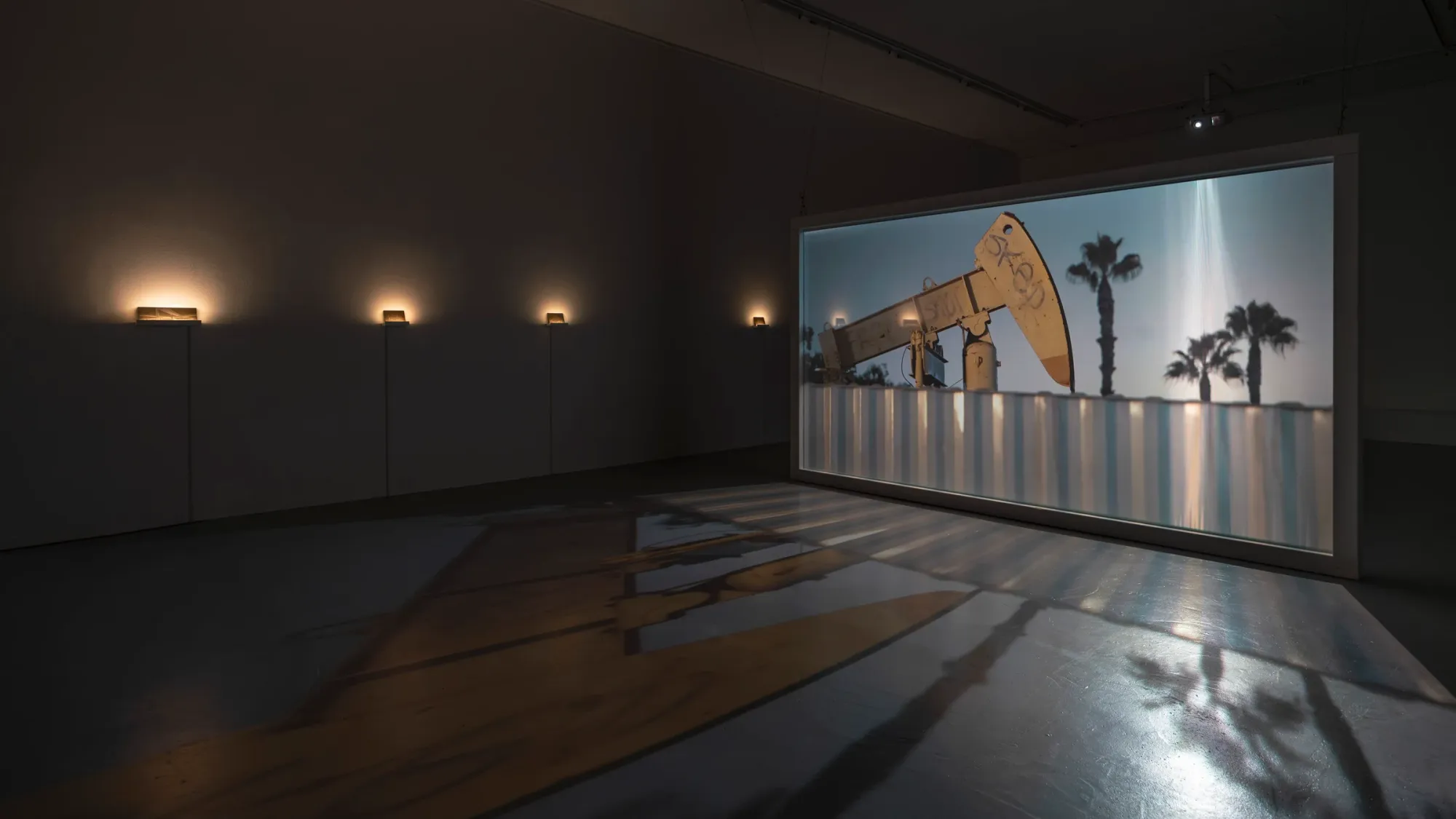 Large illuminated video installation showing oil pump jack and palm trees in dark gallery, warm wall lights and strong shadows cast across reflective floor, Kaya & Blank Crude Aesthetics basis Frankfurt 2026 exhibition view