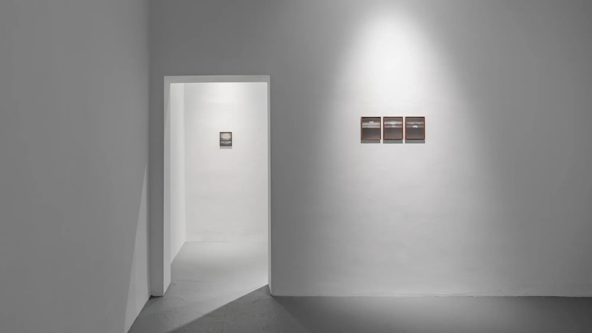Minimal gallery space with doorway leading to small framed seascape photograph, three additional framed works aligned on adjacent wall, strong light creating shadow across floor, Kaya & Blank Intermodal basis Frankfurt