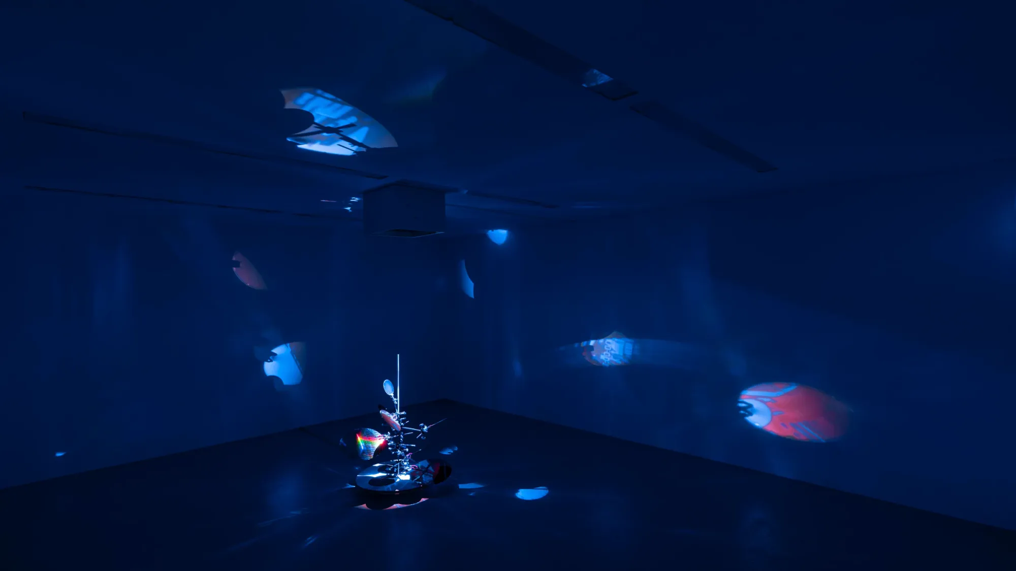 Dark immersive installation with blue lighting, reflective kinetic sculpture casting colored light projections across walls and ceiling, Kaya & Blank Stream basis Frankfurt 2026 exhibition view