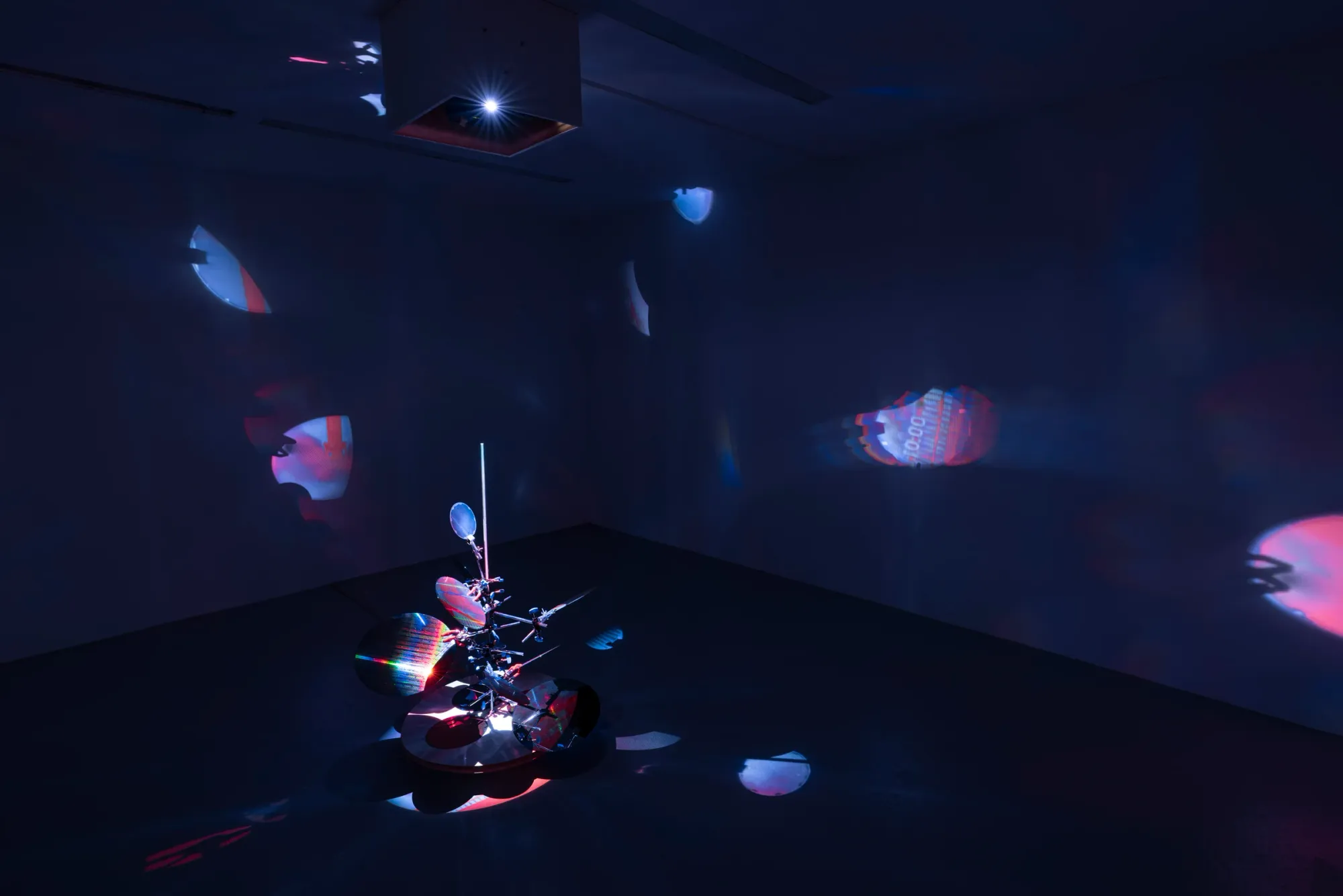 Installation view showing ceiling-mounted projector and reflective sculpture casting fragmented red and blue light projections across gallery space, Kaya & Blank Stream basis Frankfurt 2026