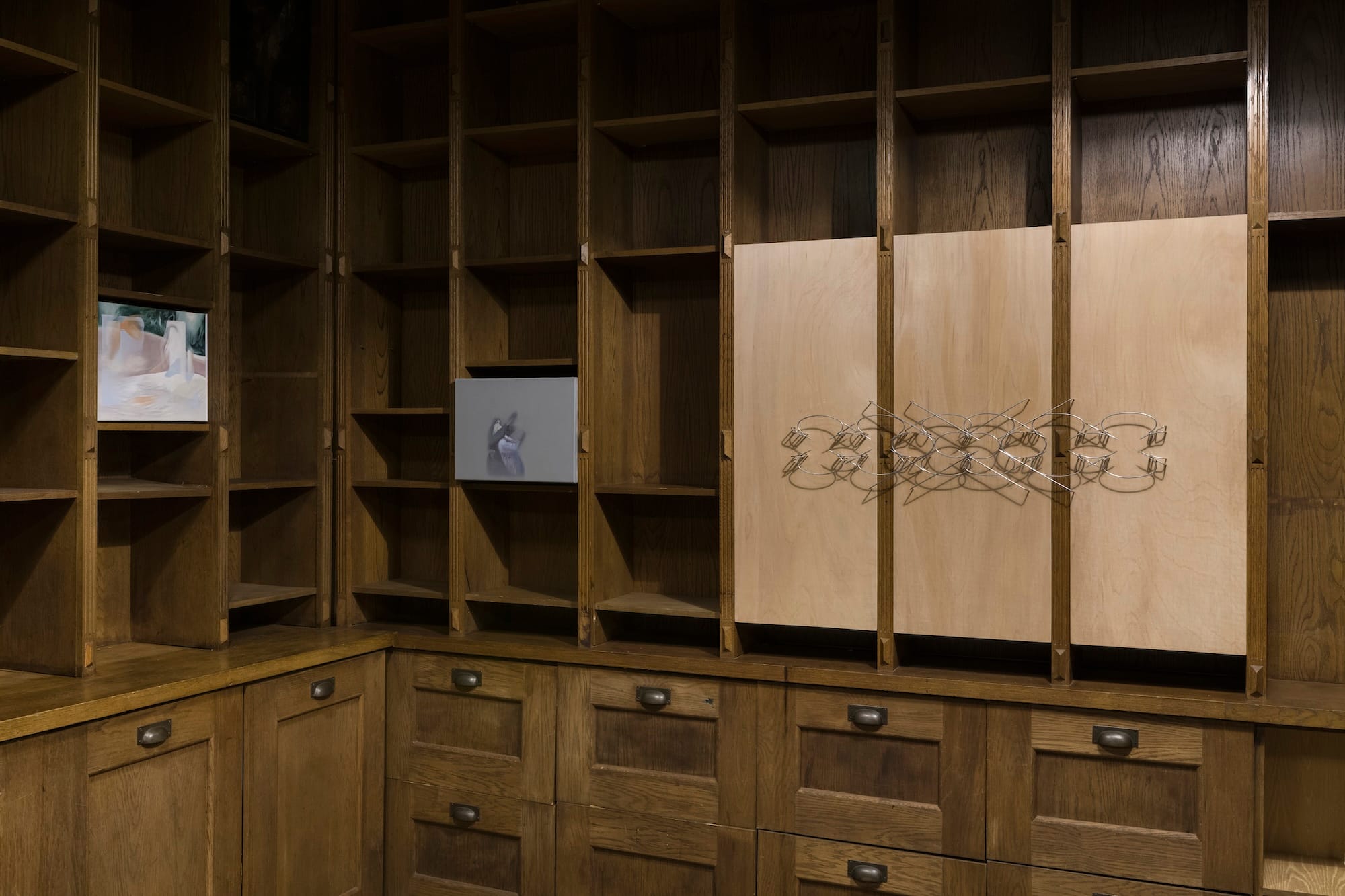 Installation view of the group exhibition Bitter Spring at MARA Projects, London, showing paintings by Bora Baboçi displayed in wooden shelving alongside a metal wall sculpture by Ben Grosse-Johannboecke.