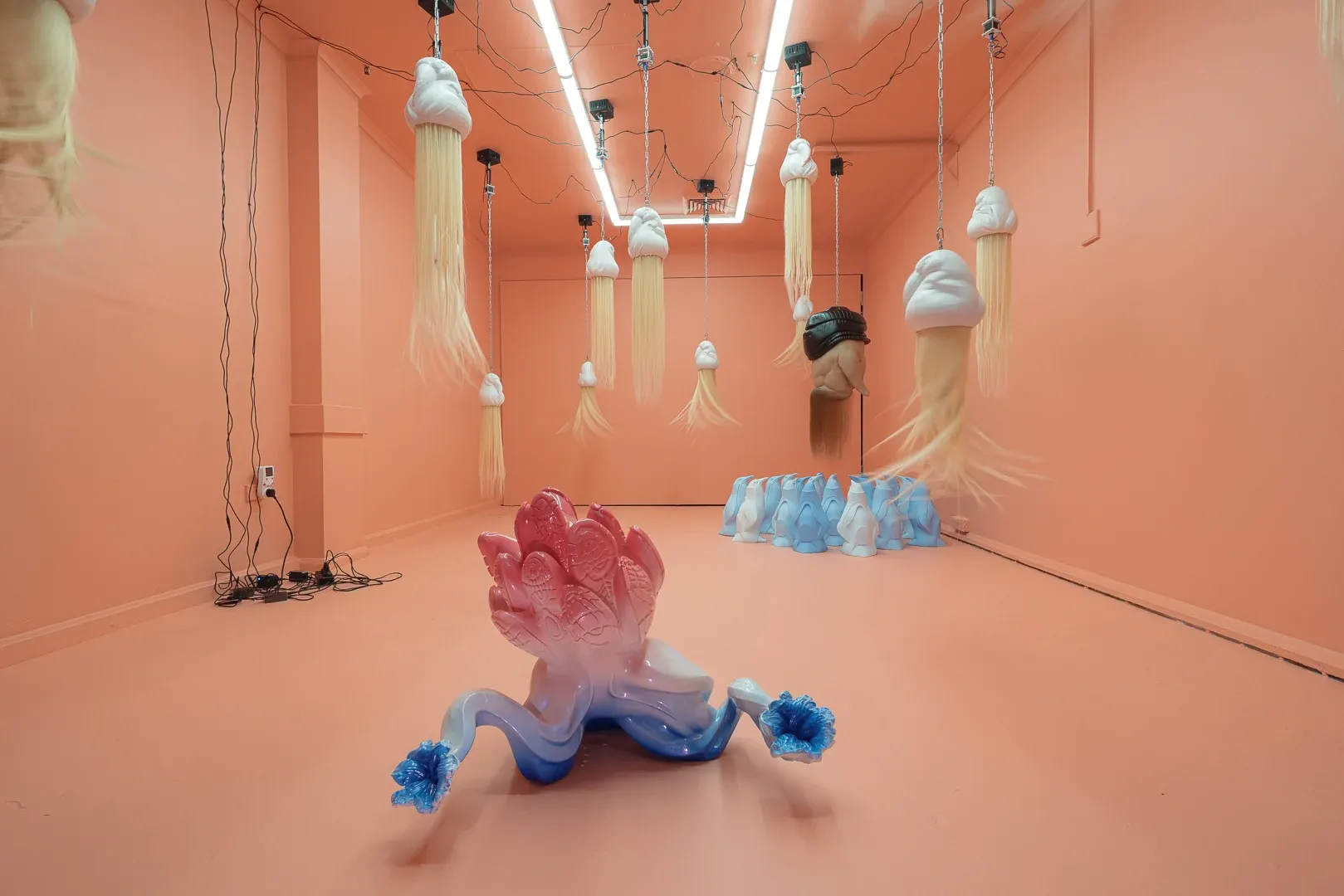 Installation view of Patricia Piccinini at Passage Gallery Sydney showing hanging kinetic hair sculptures with foreground biomorphic sculpture and blue penguin group in pink exhibition space