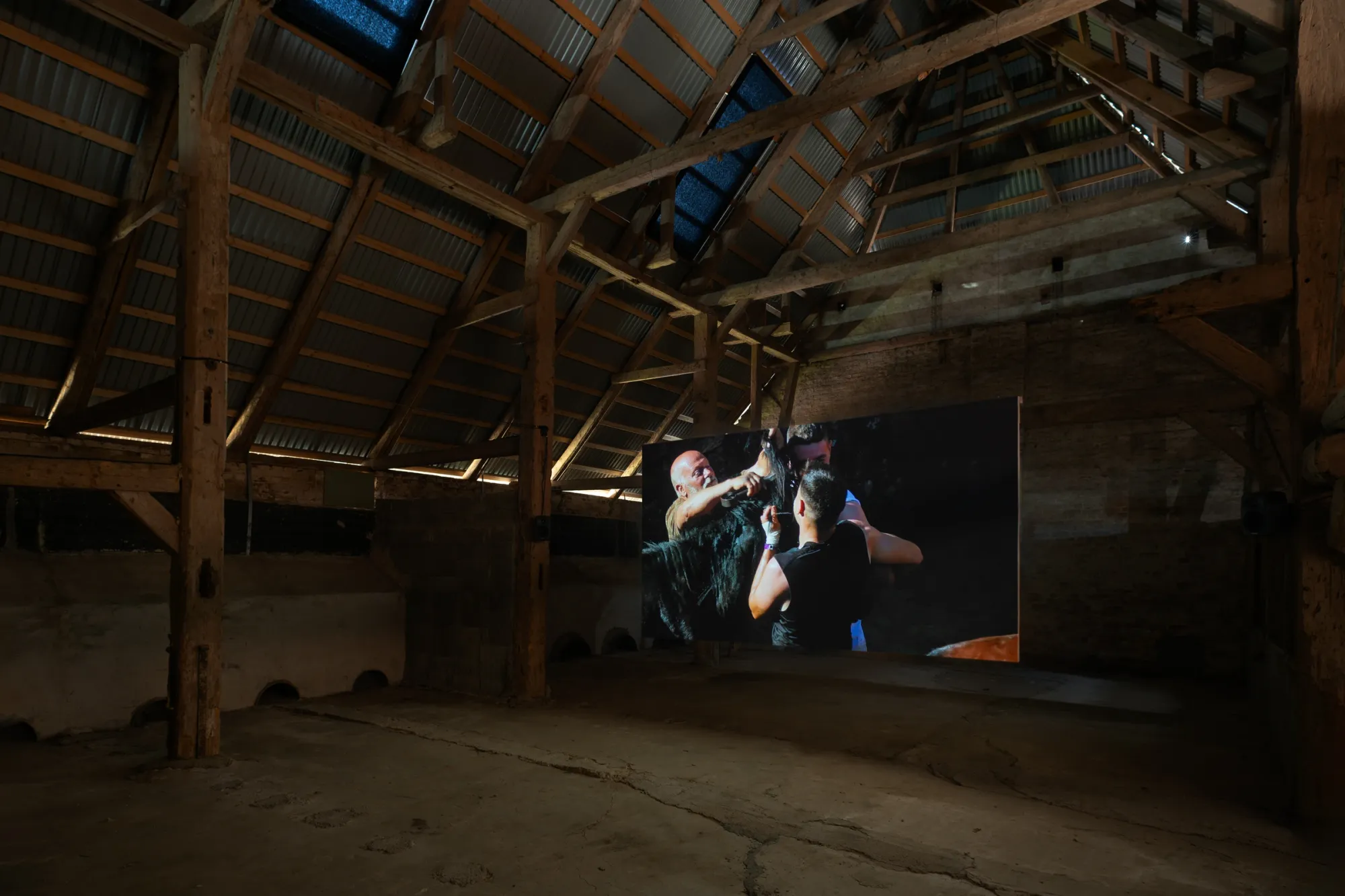 Installation view of Yalda Afsah’s exhibition Surge at Kunsthal Thy, Denmark, showing a large video projection of people handling a horse inside a wooden barn with exposed beams and concrete floor.