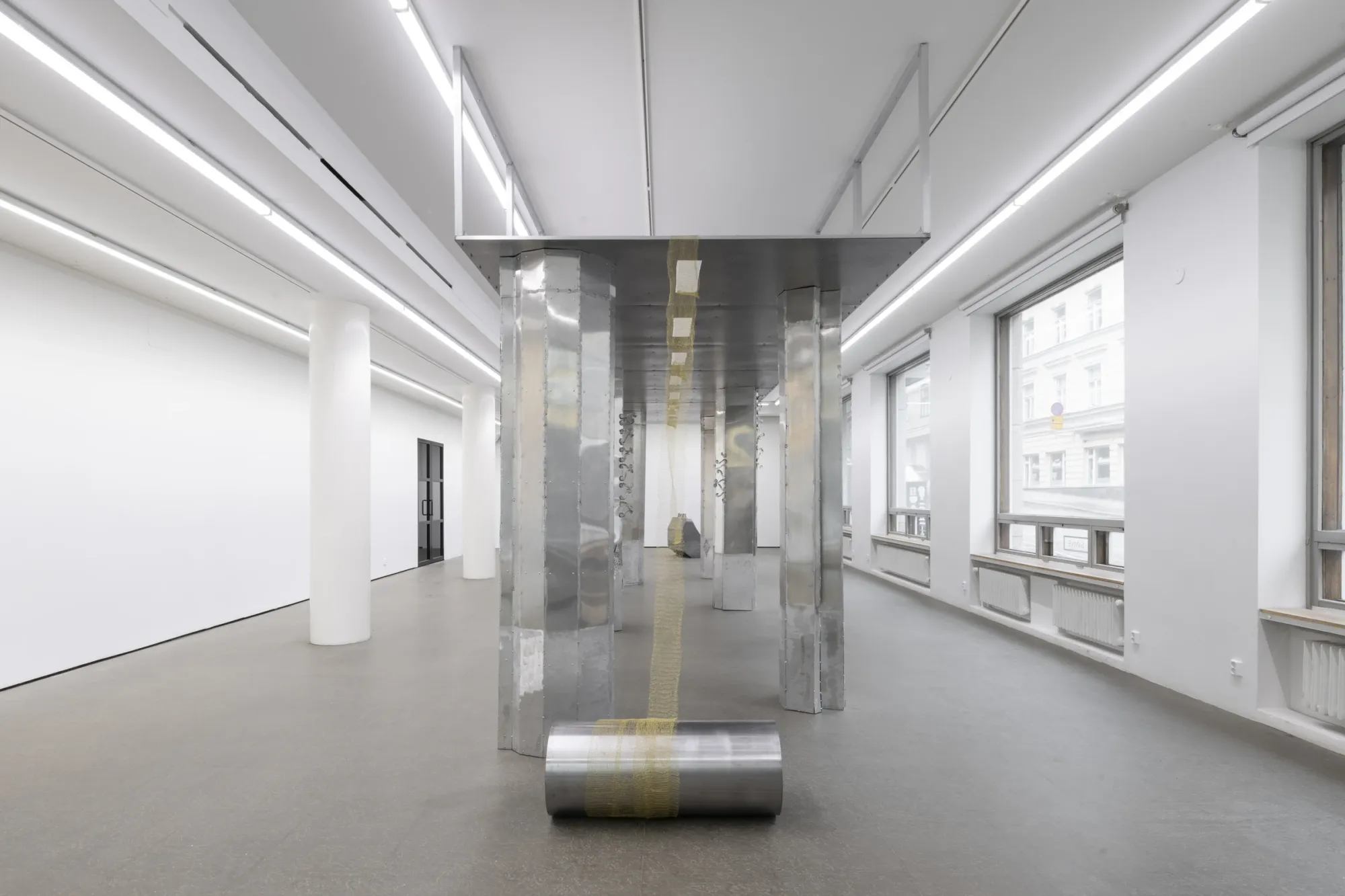  Maija Fox Hard Shoulders installation view aligned along central axis showing metal columns, suspended woven wire, and steel cylinder element in gallery