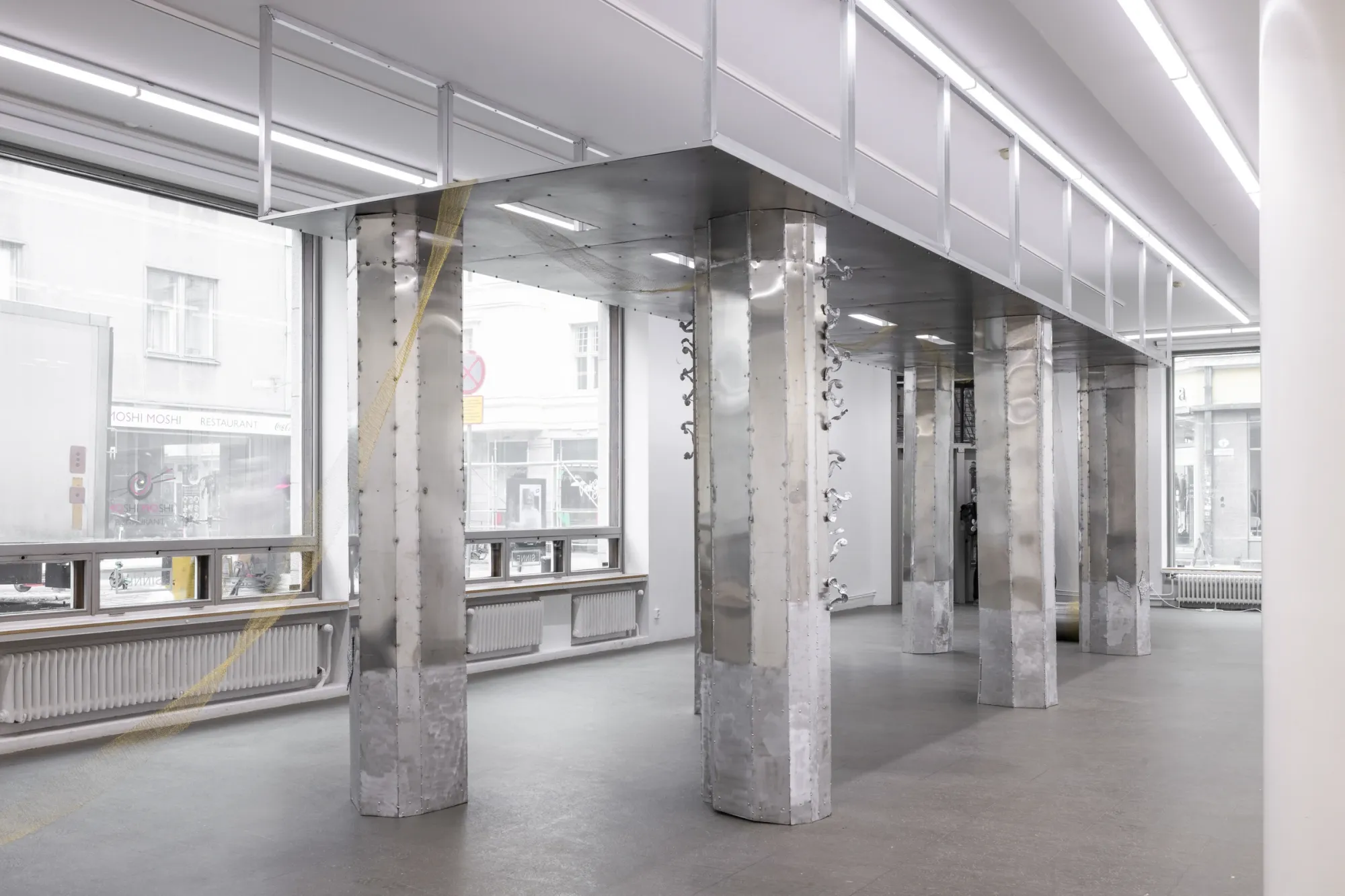 Maija Fox Hard Shoulders installation view showing metal columns and bridge-like structure with woven wire element extending toward window-lit gallery space