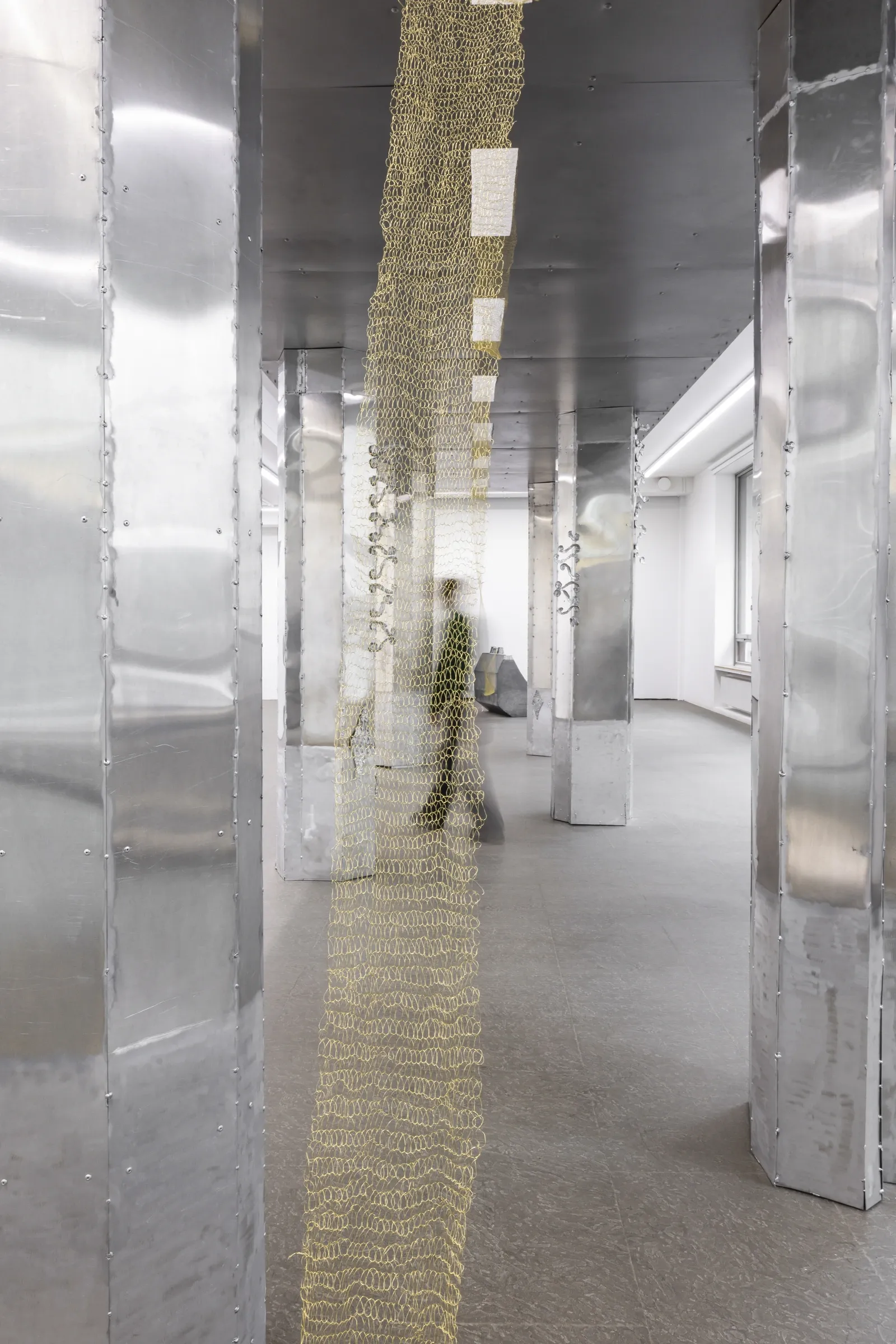 Maija Fox Hard Shoulders installation view showing woven metal wire textile with blurred human figure for scale between metal columns