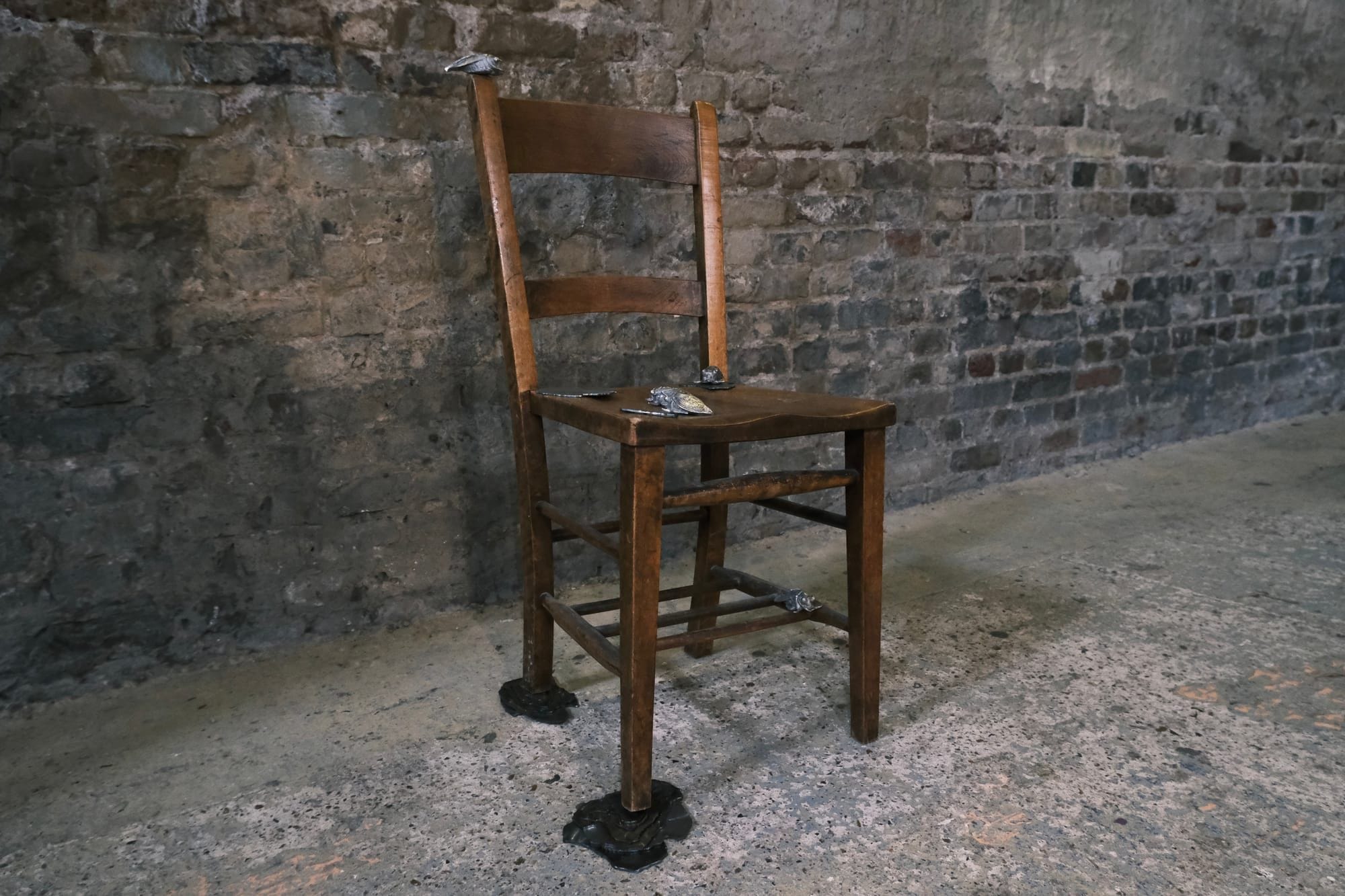 Occupied seat 2026 by Shirley Fu, wooden chair with pewter and steel elements placed on a concrete floor against a brick wall in Asylum Chapel