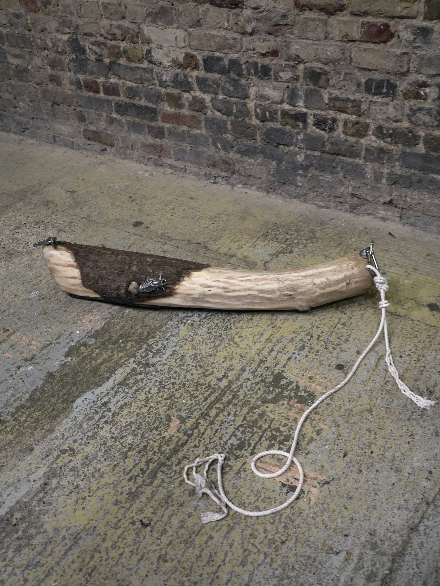 What the current left behind 2025 by Shirley Fu, sculptural work with a carved log, pewter and steel elements, and wool string placed on a concrete floor against a brick wall