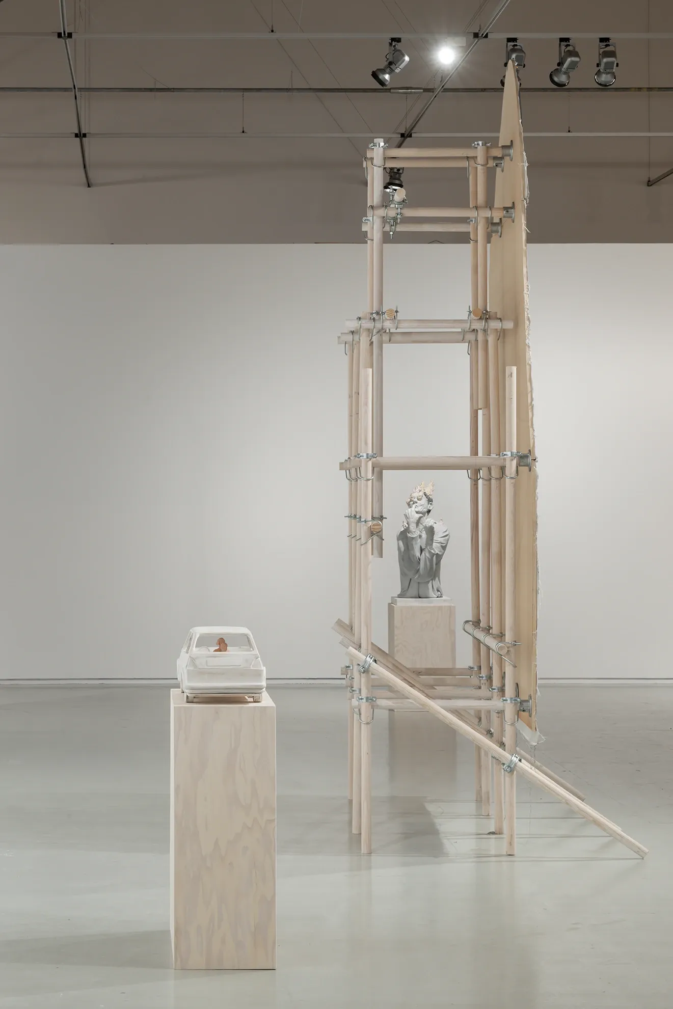 Joan Pallé, Els joves infeliços / Unhappy Youth, detail: tall arched wooden scaffolding structure enclosing a classical-style plaster bust on a plinth; white VW Golf sculpture with crouching figure on a separate plinth in the foreground left, Centre d'Art La Panera, Lleida, 2026.