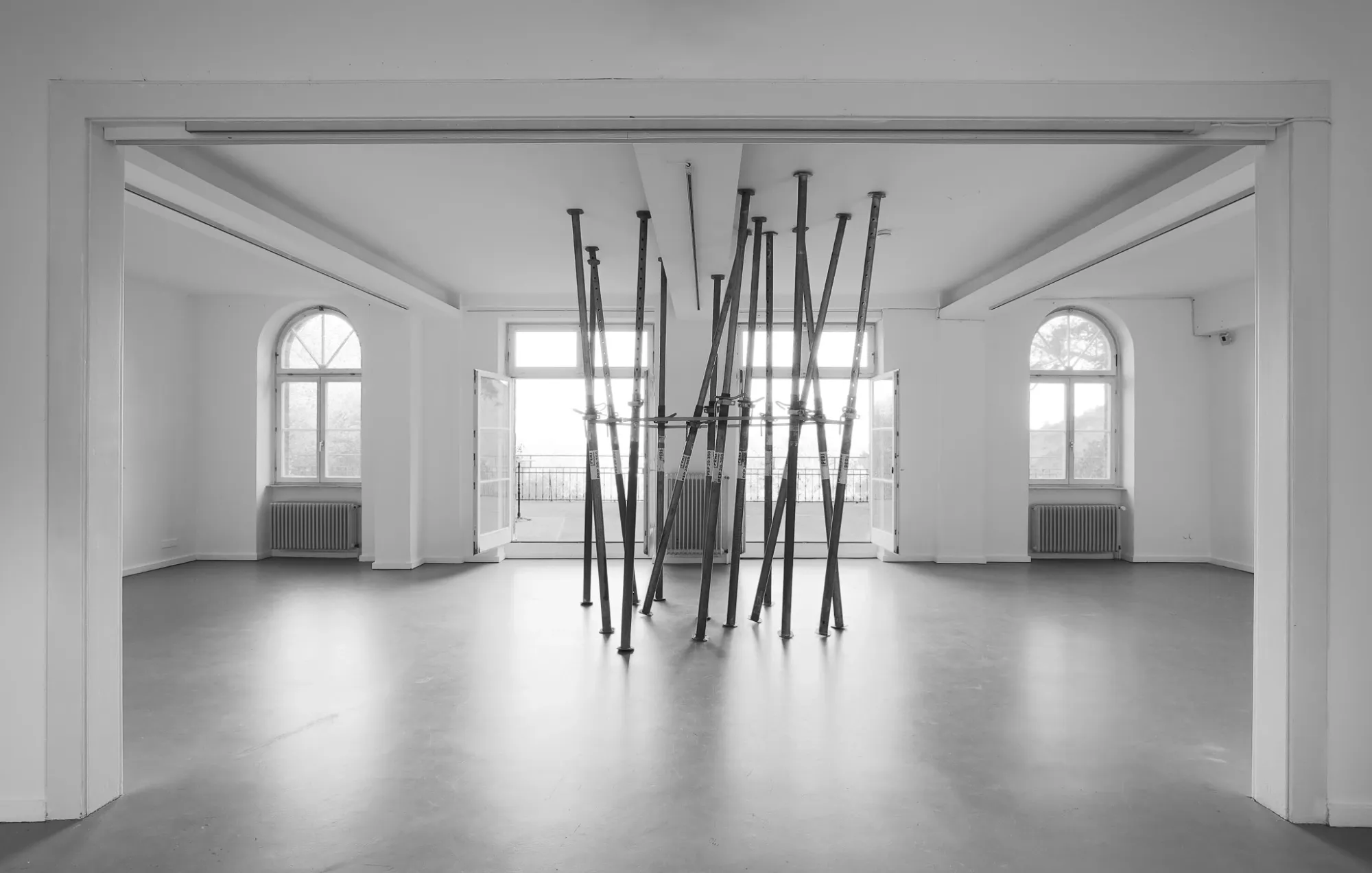 Installation view of Finja Sander's Will groß sein, will siegen at Kunstverein Wilhelmshöhe, Kassel, 2026. A single ceiling support lies horizontally on the floor, orange ratchet strap visible, two styrofoam arch shapes lean against the white wall.