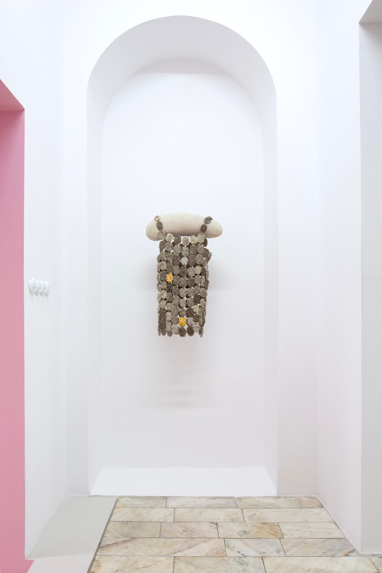 Agata Jarosławiec, We Speak in Echoes, installation view: sculptural chain-mesh curtain of grey cast silicone oval pieces linked with metal rings, suspended from a linen-covered cylindrical form, installed in a white arched niche; two gold pieces visible among the uniform grey, pink corridor wall partially visible to the left, Fundacja Galerii Piana, Kraków, 2026.