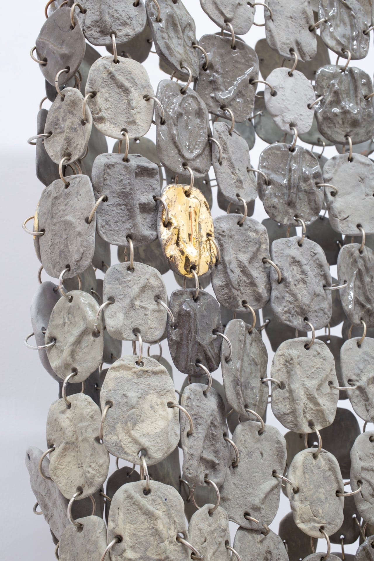 Agata Jarosławiec, We Speak in Echoes, detail: close-up of chain-mesh sculpture — hundreds of grey cast silicone oval pieces linked by metal rings, one single gold-glazed piece among them, Fundacja Galerii Piana, Kraków, 2026.