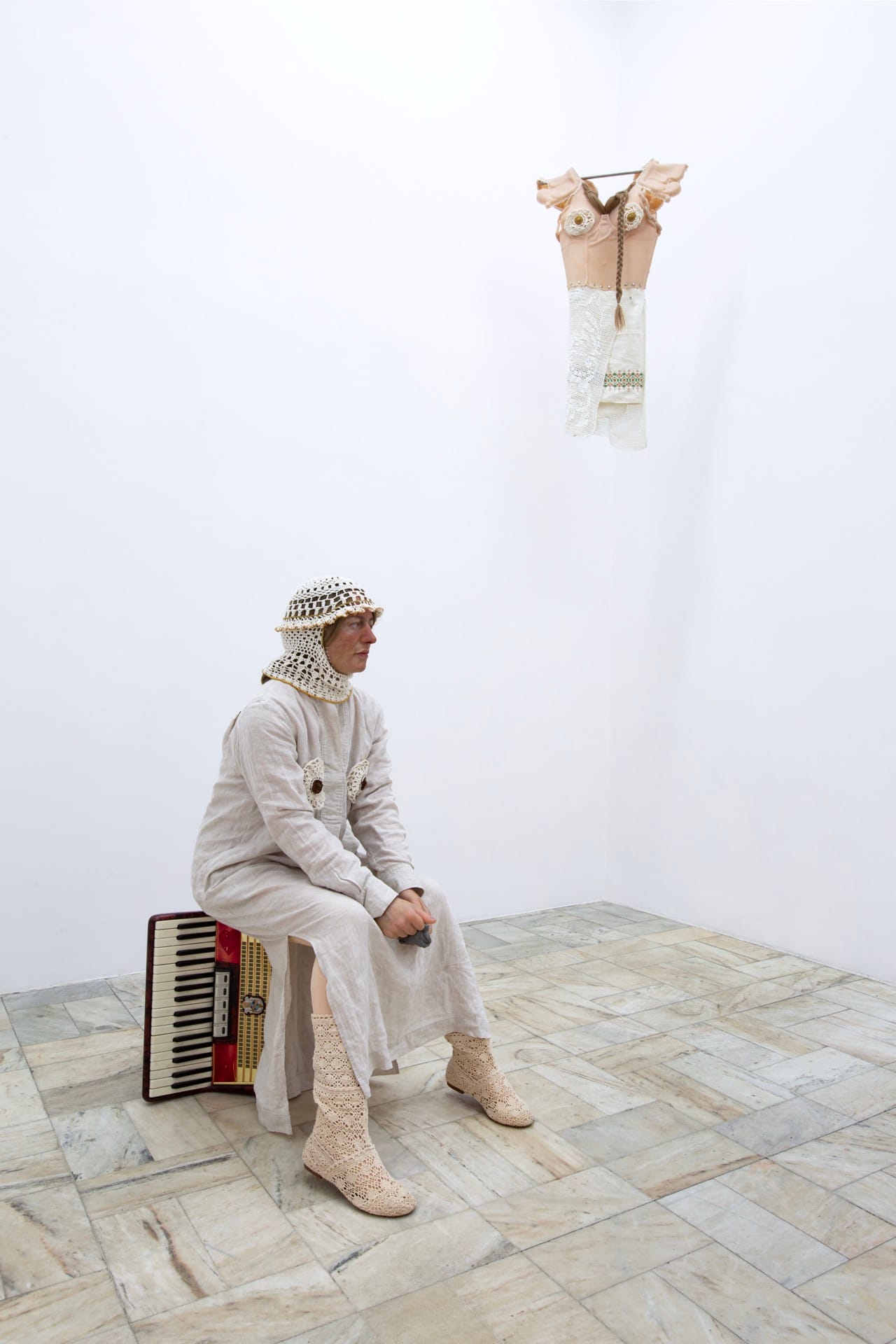 Agata Jarosławiec, We Speak in Echoes, installation view: figure in white linen suit and crocheted balaclava seated beside a vintage red accordion, suspended silicone and lace garment assemblage hanging above, Fundacja Galerii Piana, Kraków