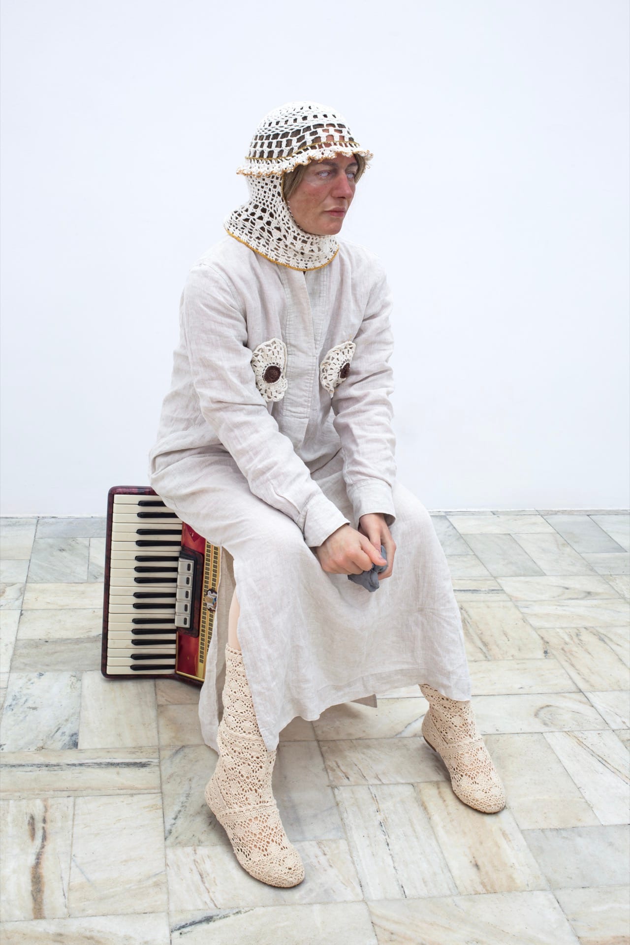 Agata Jarosławiec, We Speak in Echoes, installation view: full-length figure in white linen suit and crocheted balaclava-helmet seated on a vintage red accordion, holding a grey silicone fragment in both hands, Fundacja Galerii Piana, Kraków, 2026.