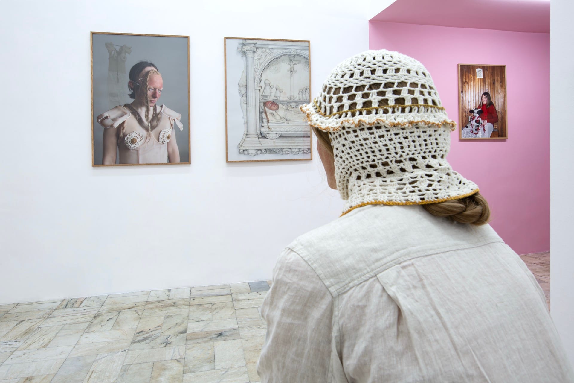 Agata Jarosławiec, We Speak in Echoes, installation view: figure in crocheted balaclava seen from behind facing three framed works — a photograph of a figure wearing a silicone bodice, a Renaissance tomb drawing, and a domestic photograph on the pink corridor wall, Fundacja Galerii Piana, Kraków,