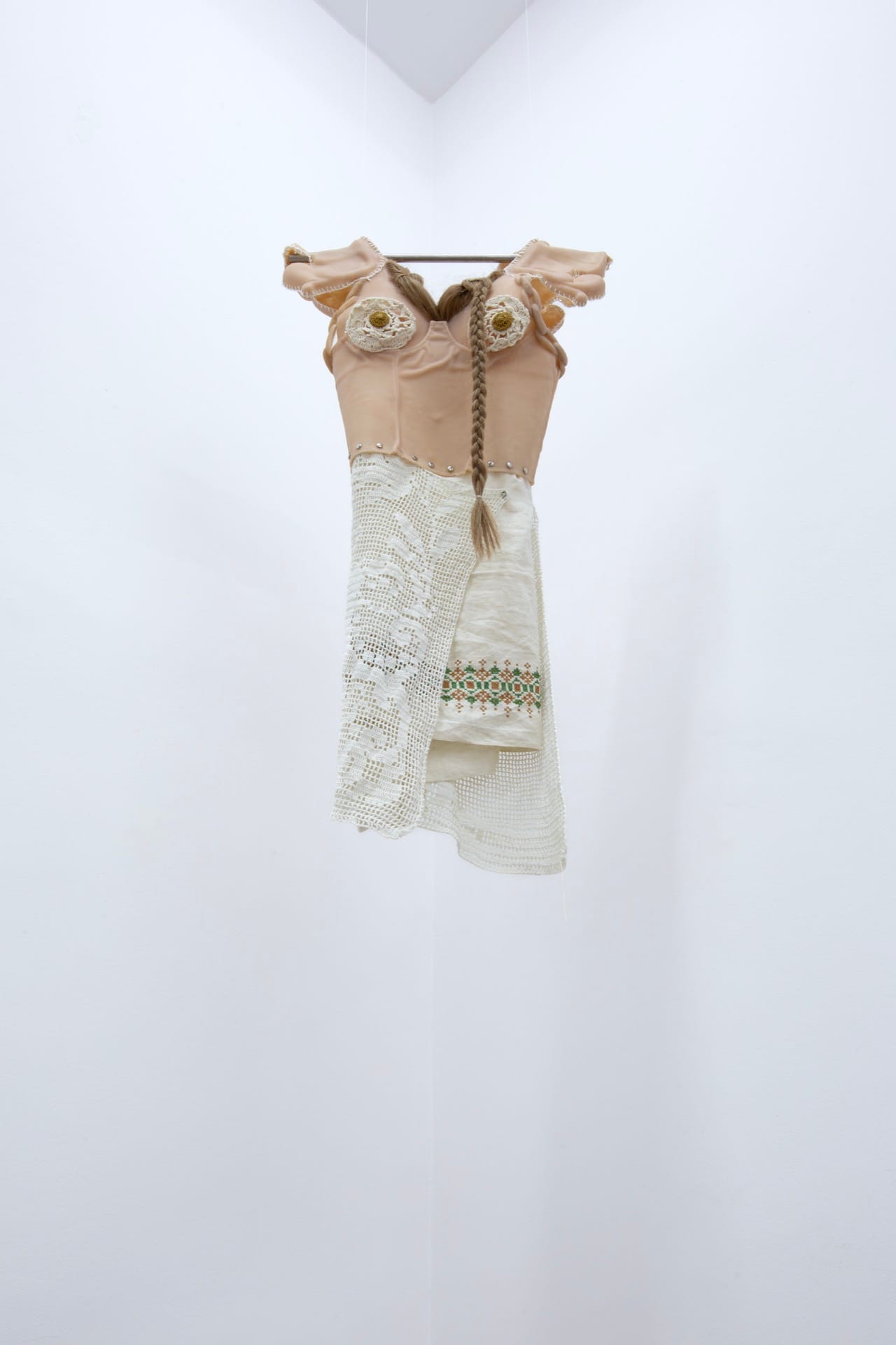 Agata Jarosławiec, We Speak in Echoes, detail: suspended garment assemblage of skin-toned silicone torso with crocheted ornaments, braided hair, folk cross-stitch embroidery in red and green, and layered lace hanging from a metal rod, Fundacja Galerii Piana, Kraków, 2026.