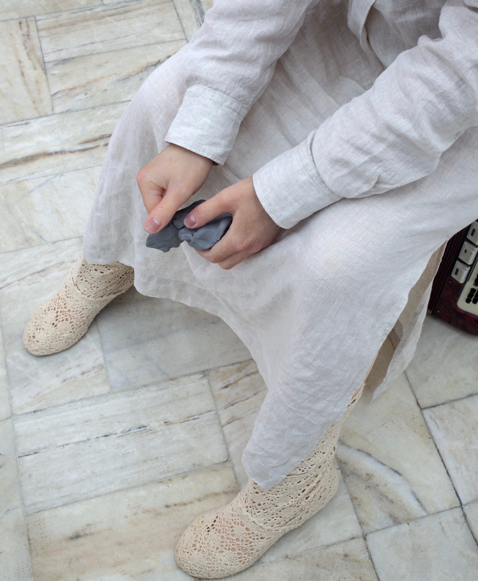 Agata Jarosławiec, We Speak in Echoes, detail: hands holding a small grey silicone fragment, crocheted lace boots on marble tile floor, accordion partially visible at right, Fundacja Galerii Piana, Kraków, 2026.