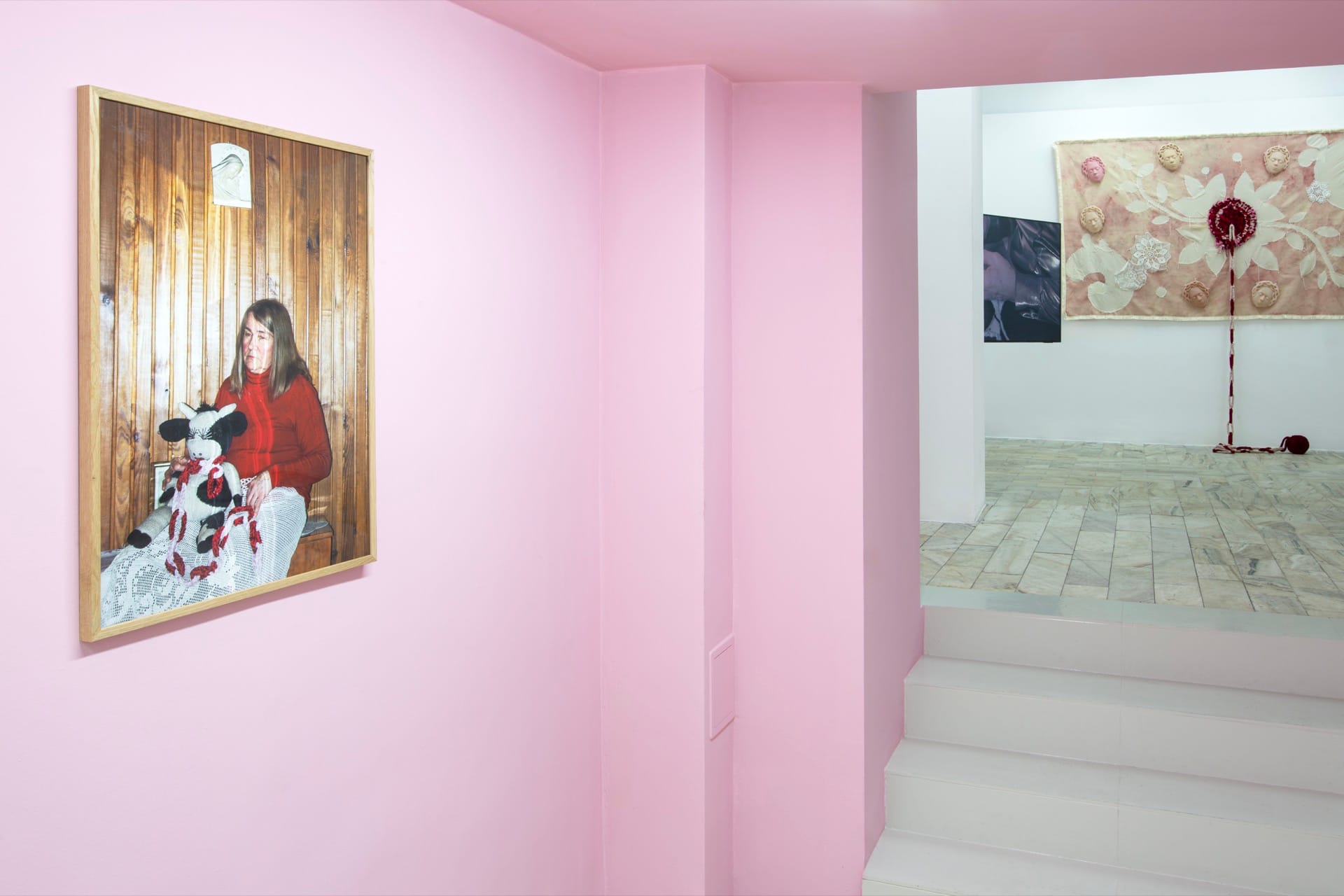 Agata Jarosławiec, We Speak in Echoes, installation view: pink-painted staircase corridor with framed domestic photograph of a woman in a red sweater holding a crocheted toy on the left wall; upper gallery visible through the opening with large textile banner and red crocheted installation, 