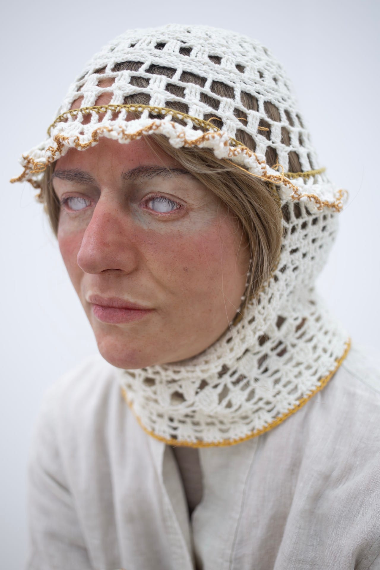 Agata Jarosławiec, We Speak in Echoes, detail: close-up portrait of figure wearing white crocheted balaclava-helmet with gold-thread trim encasing head and neck, eyes whitened with contact lenses, linen collar visible below, Fundacja Galerii Piana, Kraków, 2026.