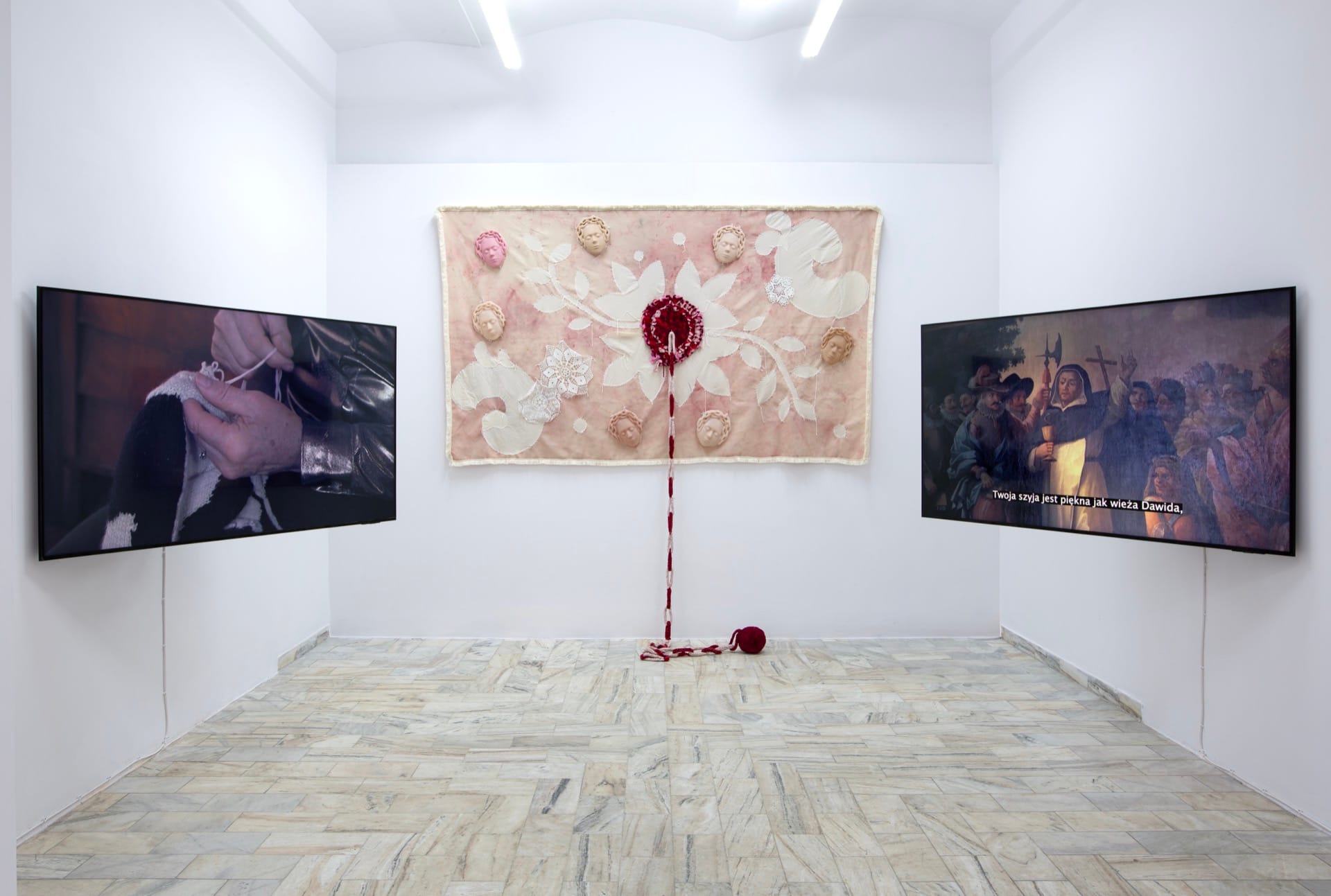 Agata Jarosławiec, We Speak in Echoes, installation view: upper gallery triptych — left video screen showing close-up of hands crocheting with dark materials; centre: large textile banner with appliquéd white flower forms, silicone face-casts, and red crocheted rose from which a braided cord descends to a ball of red yarn on the floor; right video screen showing a Polish historical religious painting with subtitle "Twoja szyja jest piękna jak wieża Dawida", Fundacja Galerii Piana, Kraków, 2026.