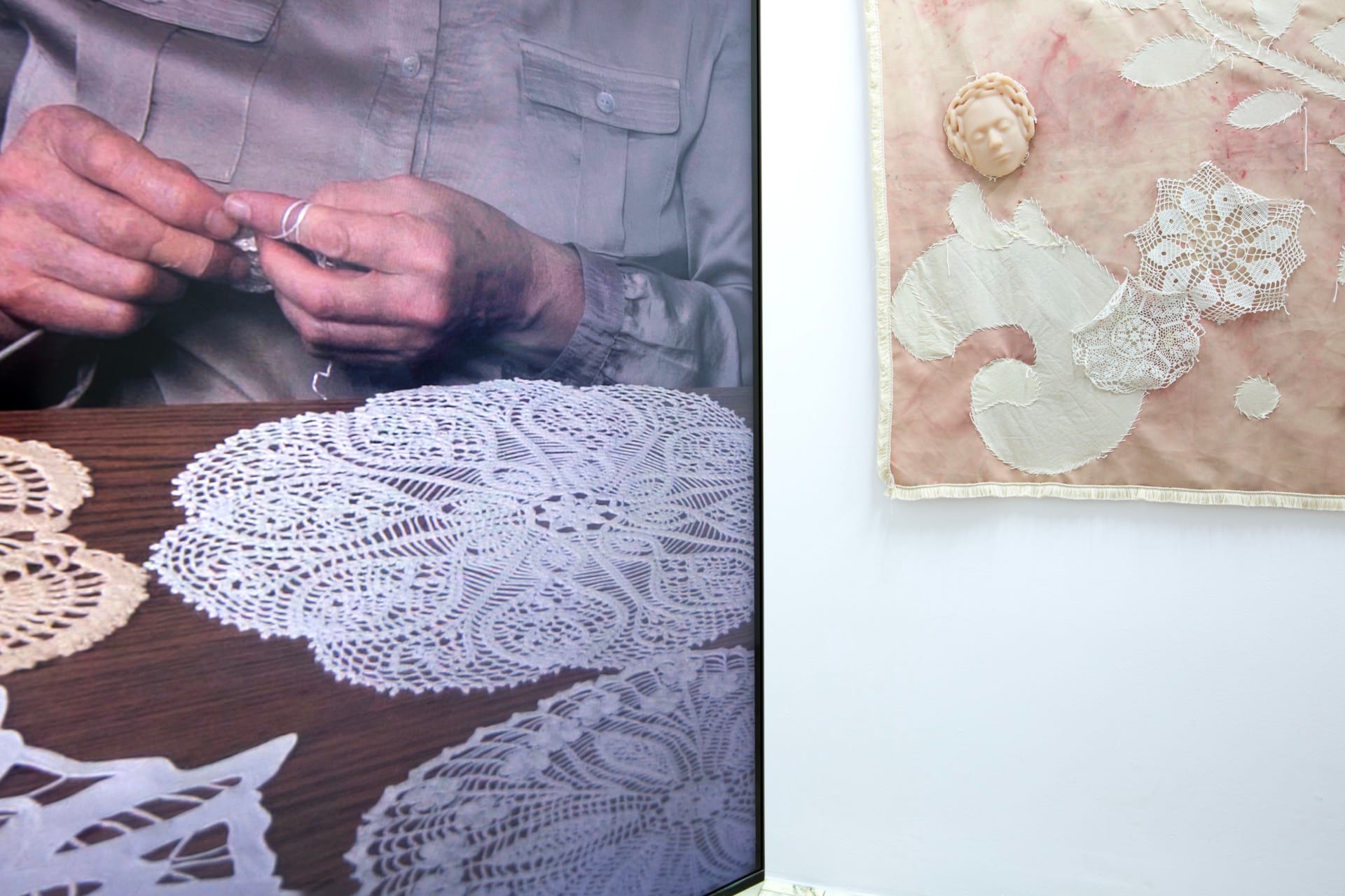 Agata Jarosławiec, We Speak in Echoes, installation detail: left video screen showing elderly hands crocheting lace doilies on a wooden table, adjacent to the large textile banner with pink-beige ground and a silicone face-cast with braided hair crown, Fundacja Galerii Piana, Kraków, 2026.