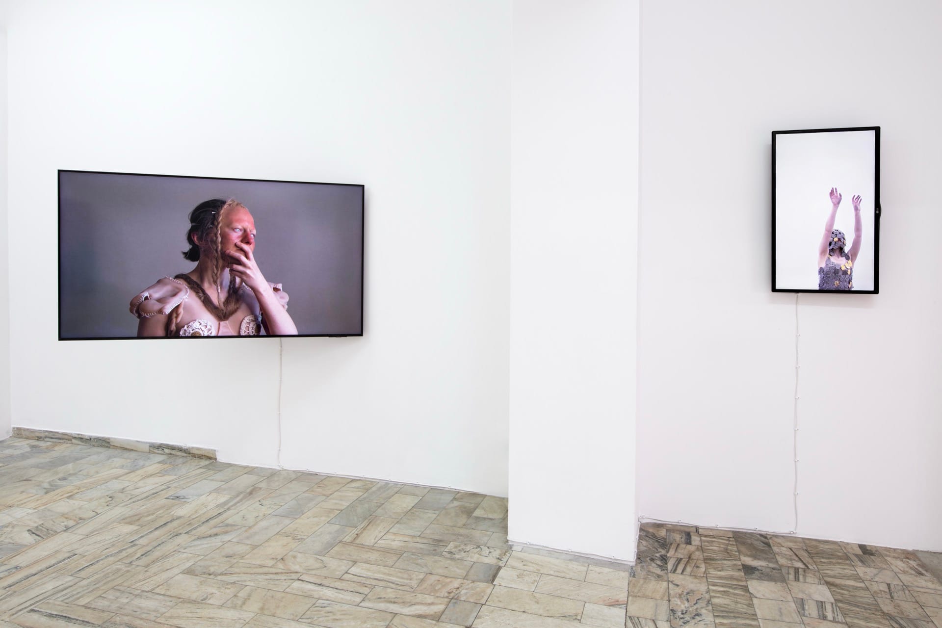Agata Jarosławiec, We Speak in Echoes, installation view: large video screen showing a figure in a skin-toned silicone bodice with crocheted ornaments touching their face; smaller vertical screen showing a figure in crocheted costume with arms raised upward against white background, Fundacja Galerii Piana, Kraków, 2026.