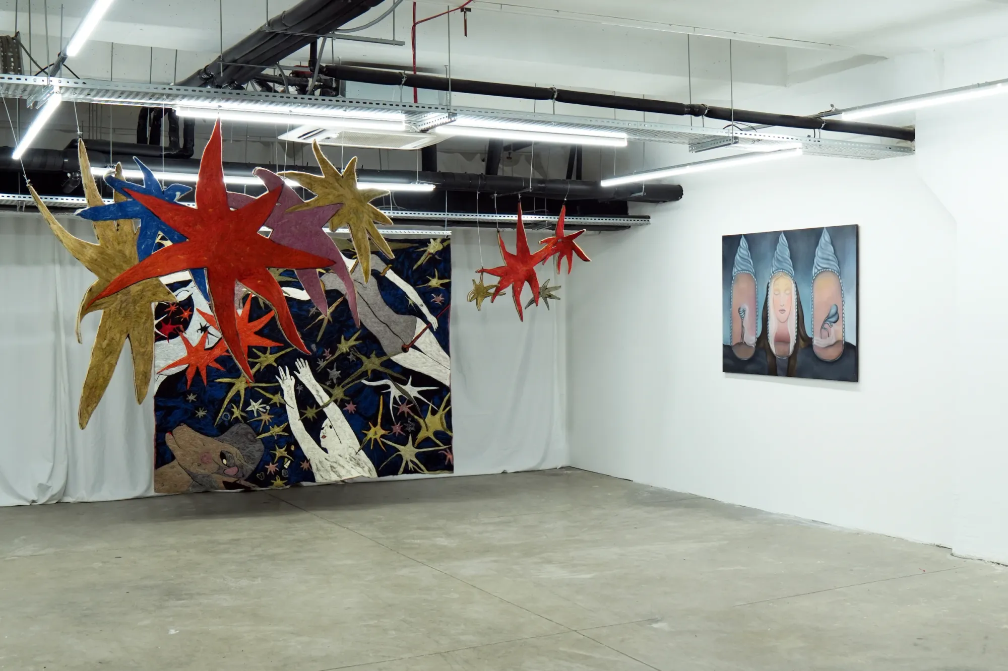 Catalogue of Sensory Data, installation view — left: Gvantsa Jishkariani, Catalogue of Sensory Data, 2026, large-scale felt tapestry with suspended star-shaped felt sculptures; right: Nata Varazi, Untitled, 2025, oil on canvas depicting two large face forms with conical hats and birds; The Why Not Gallery at Fabrika Project Space, Tbilisi.