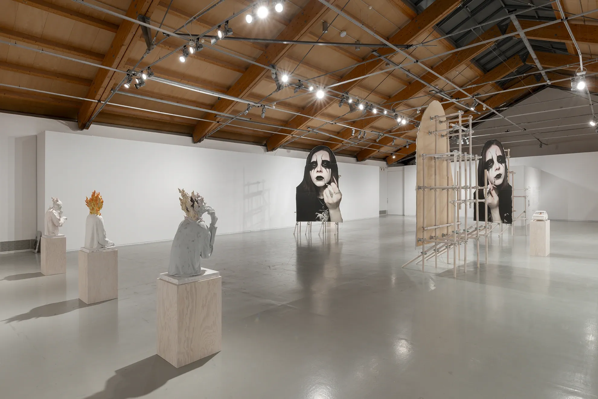 Installation view of Joan Pallé, Els joves infeliços / Unhappy Youth, Centre d'Art La Panera, Lleida, 2026. Left: two plaster figurative sculptures on wooden plinths; centre and right: monumental goth portrait photographs on easels and large arched wooden scaffolding structures; wooden car sculpture on plinth in background