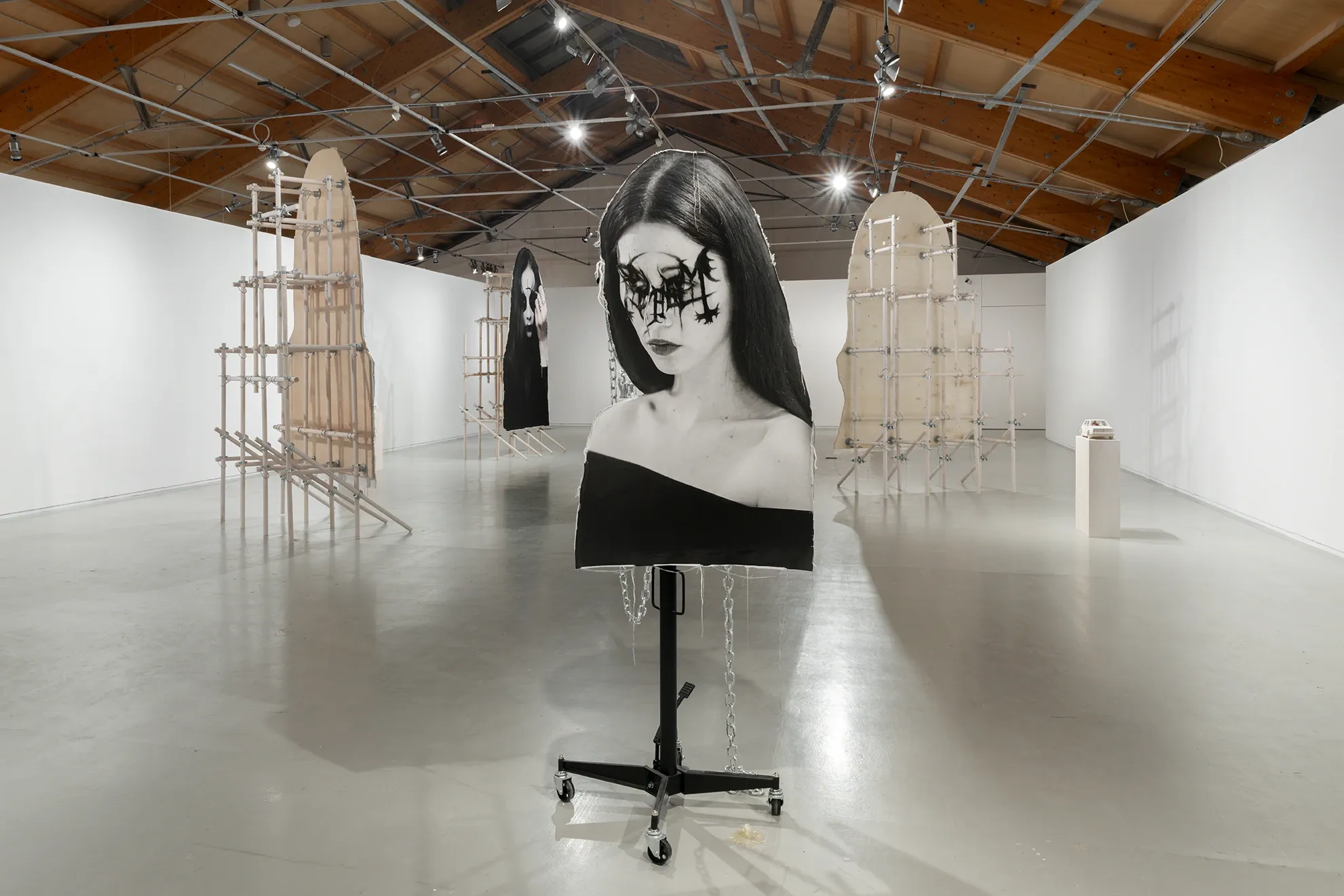 Installation view of Joan Pallé, Els joves infeliços / Unhappy Youth, Centre d'Art La Panera, Lleida, 2026. Foreground: black-and-white portrait of a young woman with elaborate goth face paint on a wheeled stand with hanging chains; arched empty wooden scaffolding frames flank the space; a second portrait visible in the background.
