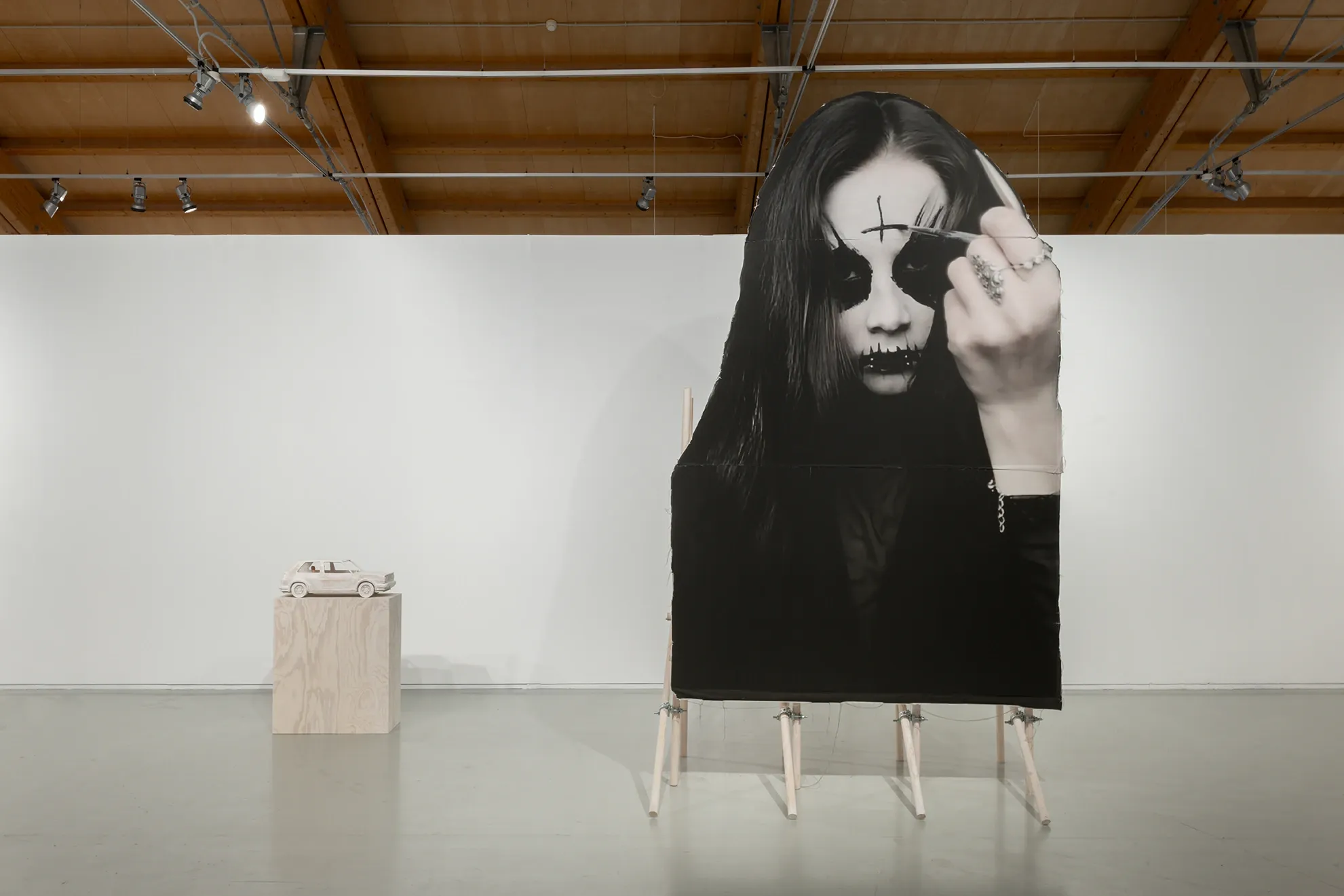 Installation view of Joan Pallé, Els joves infeliços / Unhappy Youth, Centre d'Art La Panera, Lleida, 2026. Monumental black-and-white photograph of a young woman in black metal makeup giving the middle finger, mounted on wooden easel; small wooden car sculpture on plinth in background.