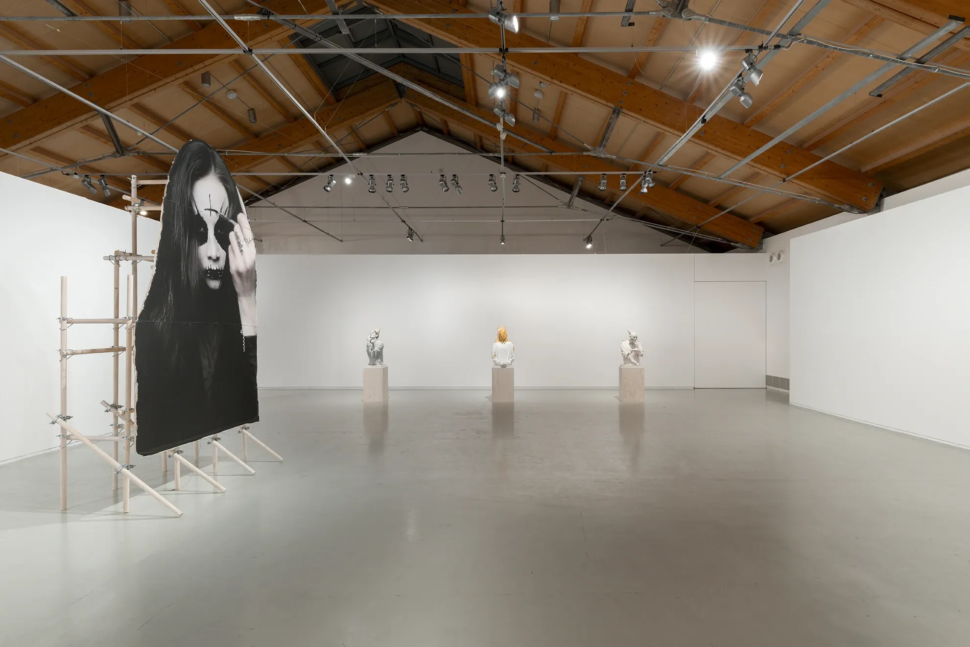 Installation view of Joan Pallé, Els joves infeliços / Unhappy Youth, Centre d'Art La Panera, Lleida, 2026. Left: monumental goth portrait photograph on wooden scaffolding; centre and right: three plaster figurative sculptures on wooden plinths in a row — a classical bust, a flame-headed sweatshirt figure, and a robed figure — seen from behind against the white gallery wall.