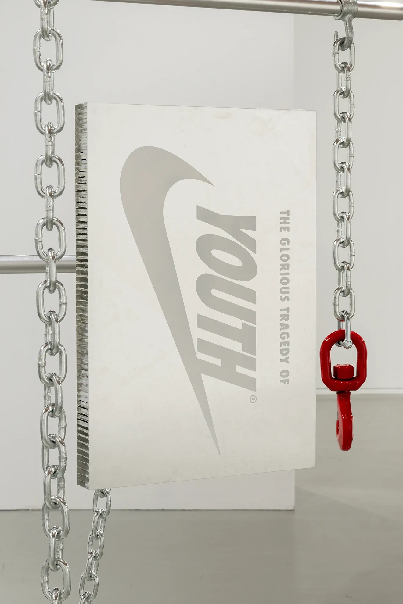 Joan Pallé, Els joves infeliços / Unhappy Youth, detail: panel reading "youth / The Glorious Tragedy Of" with a Nike-style swoosh logo, suspended by industrial chains with a red hook, Centre d'Art La Panera, Lleida, 2026.