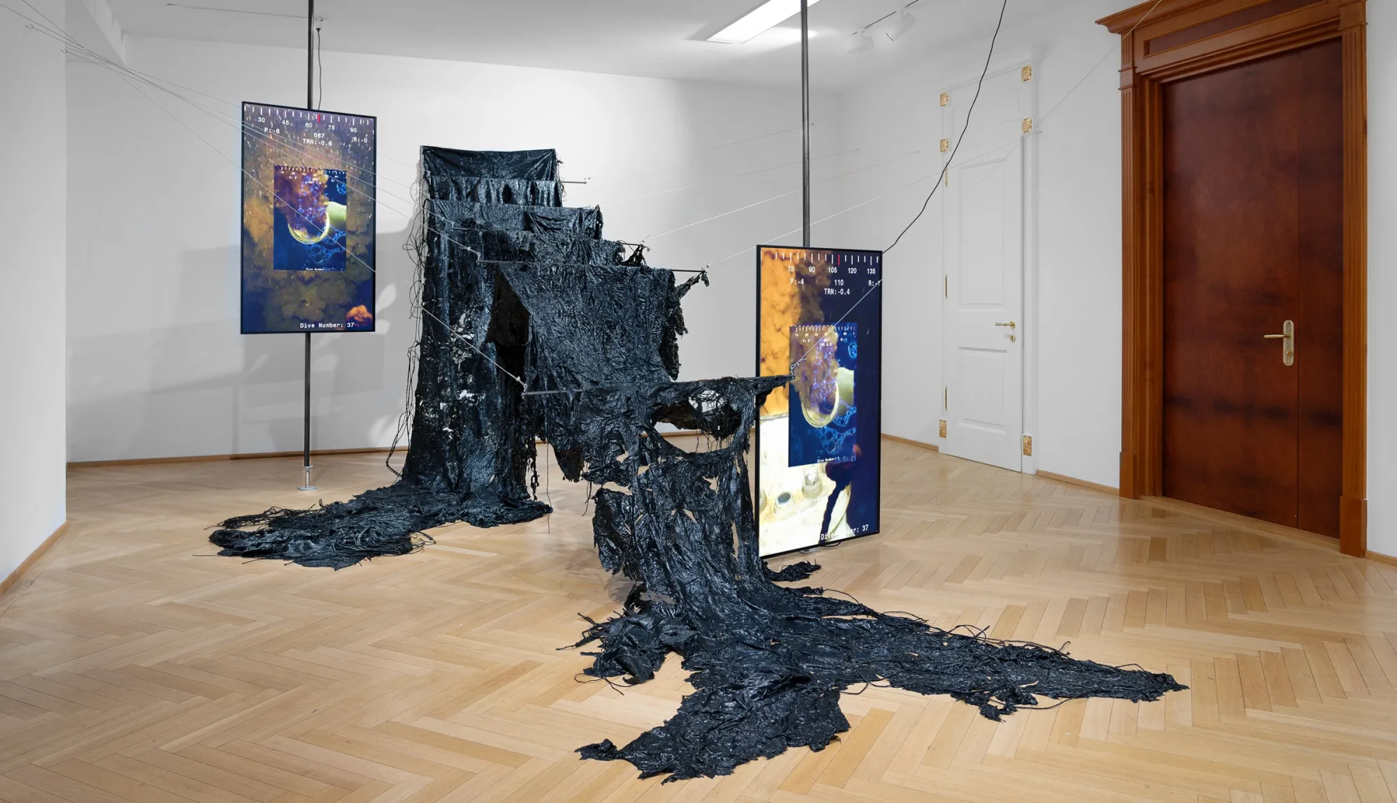 Kaja Clara Joo, 다시 : DASI, 2026, installation view. Black degraded latex suspended between two steel poles draped across parquet floor, flanked by two vertical video monitors showing deep-sea survey footage with oceanic and gold imagery. Korean Culture Center Vienna.