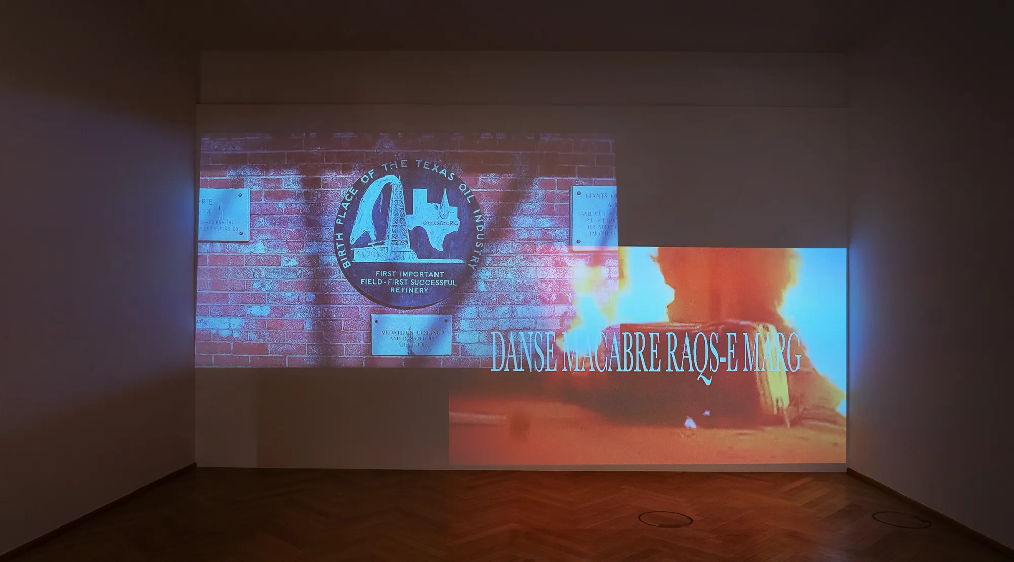 Kaja Clara Joo, Danse Macabre Raqs-e Marg, 2026, two-channel video projection, installation view. Left: commemorative plaque "Birthplace of the Texas Oil Industry – Corsicana". Right: fire footage with title text "Danse Macabre Raqs-e Marg". Korean Culture Center Vienna.