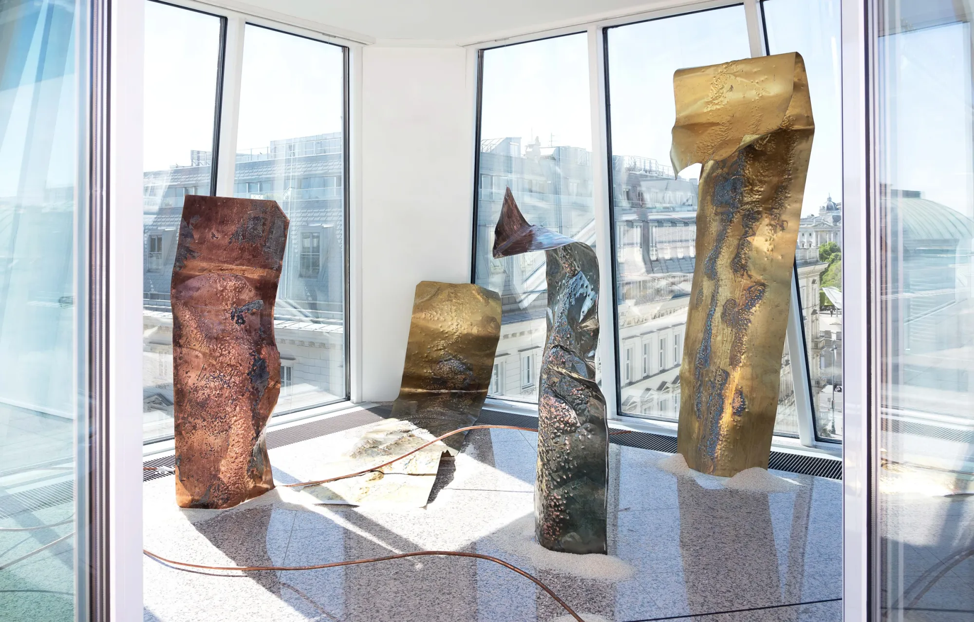 Kaja Clara Joo, 다시 : DASI, 2026, installation view. Large vertical metal slabs in copper, brass and oxidized steel standing in white sand, glass-walled rooftop pavilion with Vienna Ringstrasse panorama in background. Korean Culture Center Vienna.