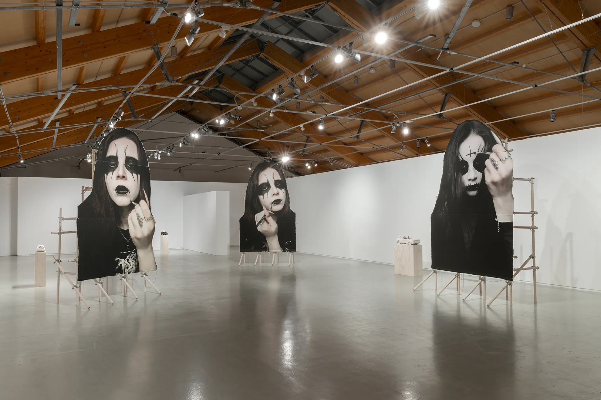 Installation view of Joan Pallé, Els joves infeliços / Unhappy Youth, Centre d'Art La Panera, Lleida, 2026. Three monumental black-and-white photographs of young women in black metal makeup giving the middle finger, mounted on wooden scaffolding; small wooden car sculptures on plinths throughout the space.