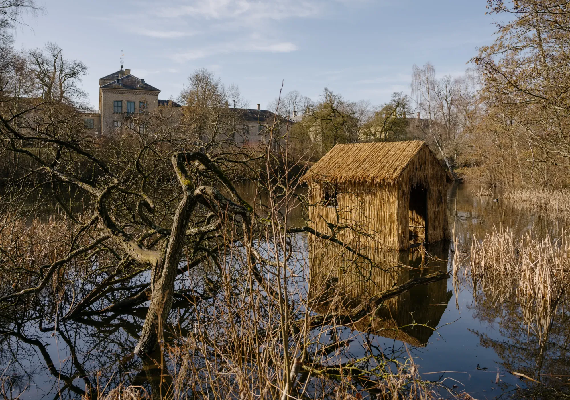 Olivia Rode Hvass, She listens but she can't speak, installation view, Museet for Samtidskunst, Roskilde — reed house floating on the still lake, framed by bare winter branches, the historic Kurhuset building visible in the background.