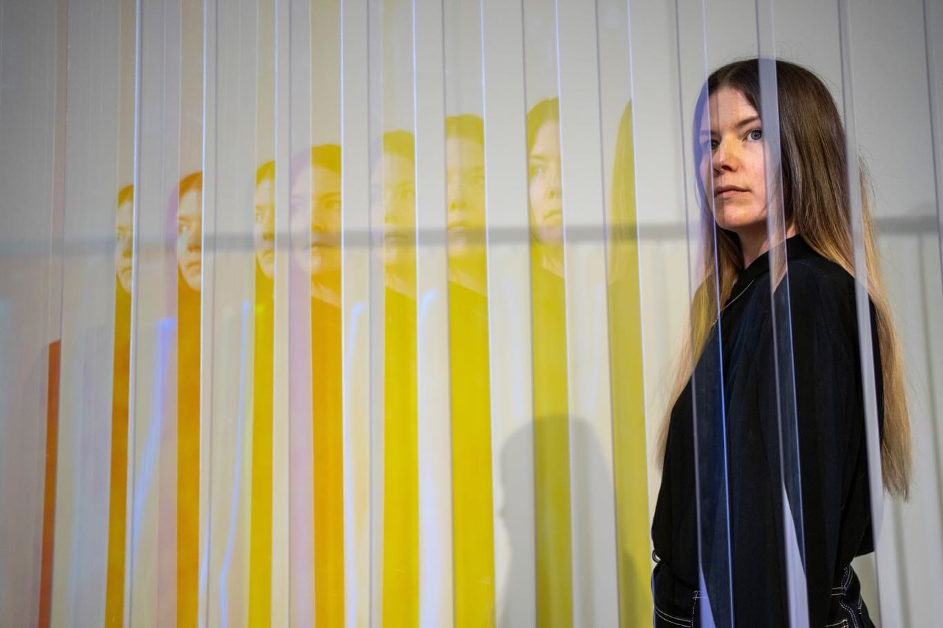 Thordis Erla Zoega inside Everyday, portrait: artist seen through vertical blind slats — her face and figure multiplied by dichroic reflections into a sequence of coloured fragments in yellow, orange, and blue, warm light to the left, BERG Contemporary, Reykjavík, 2025.