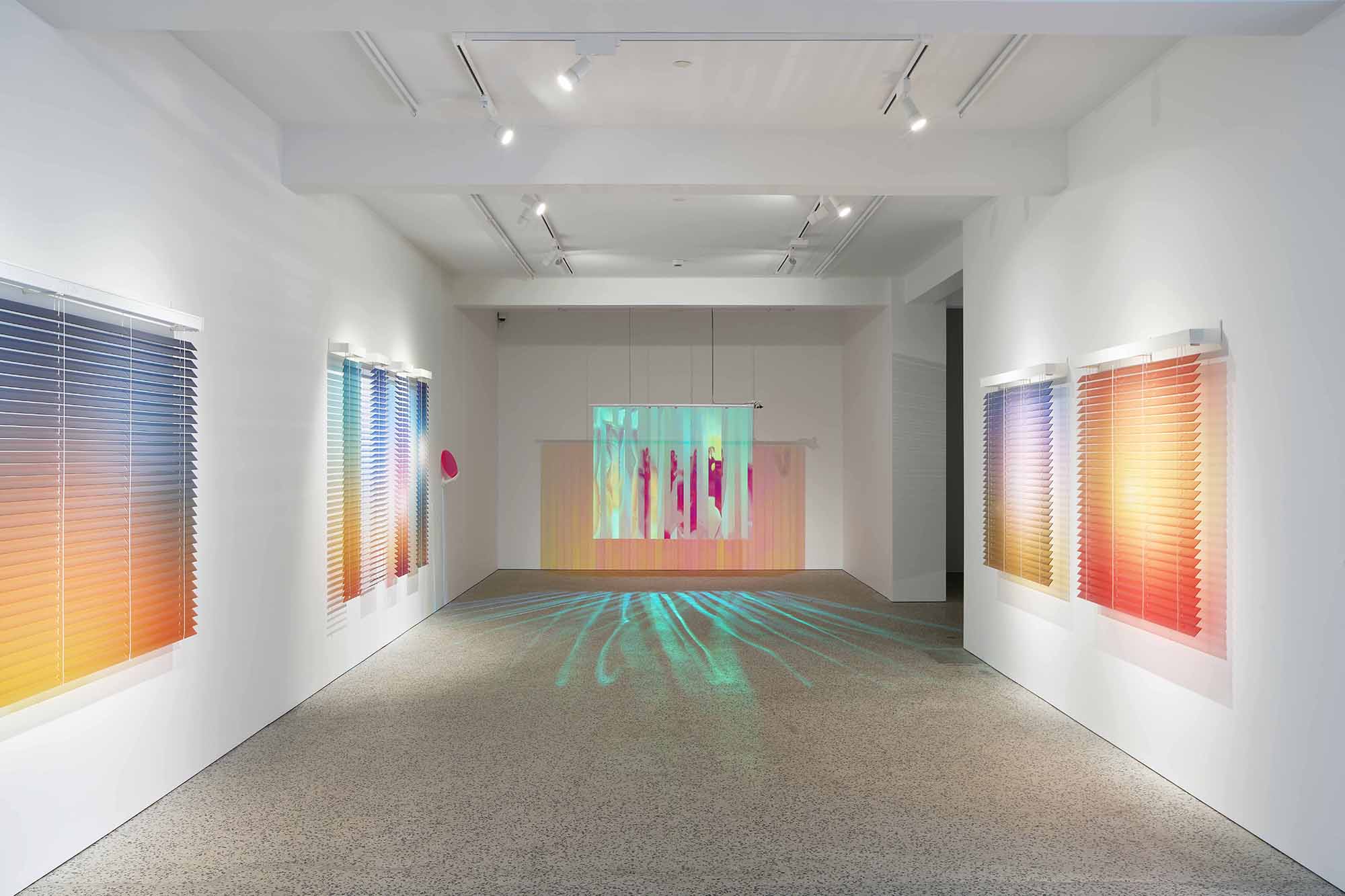 Thordis Erla Zoega, Domestic Sci-fi, installation view: four Forecast works — gradient venetian blinds running from blue-violet to warm orange — mounted on white walls left and right; Everyday installation visible at centre background with iridescent multicolour projection and teal light rays on floor, BERG Contemporary, Reykjavík, 2025.