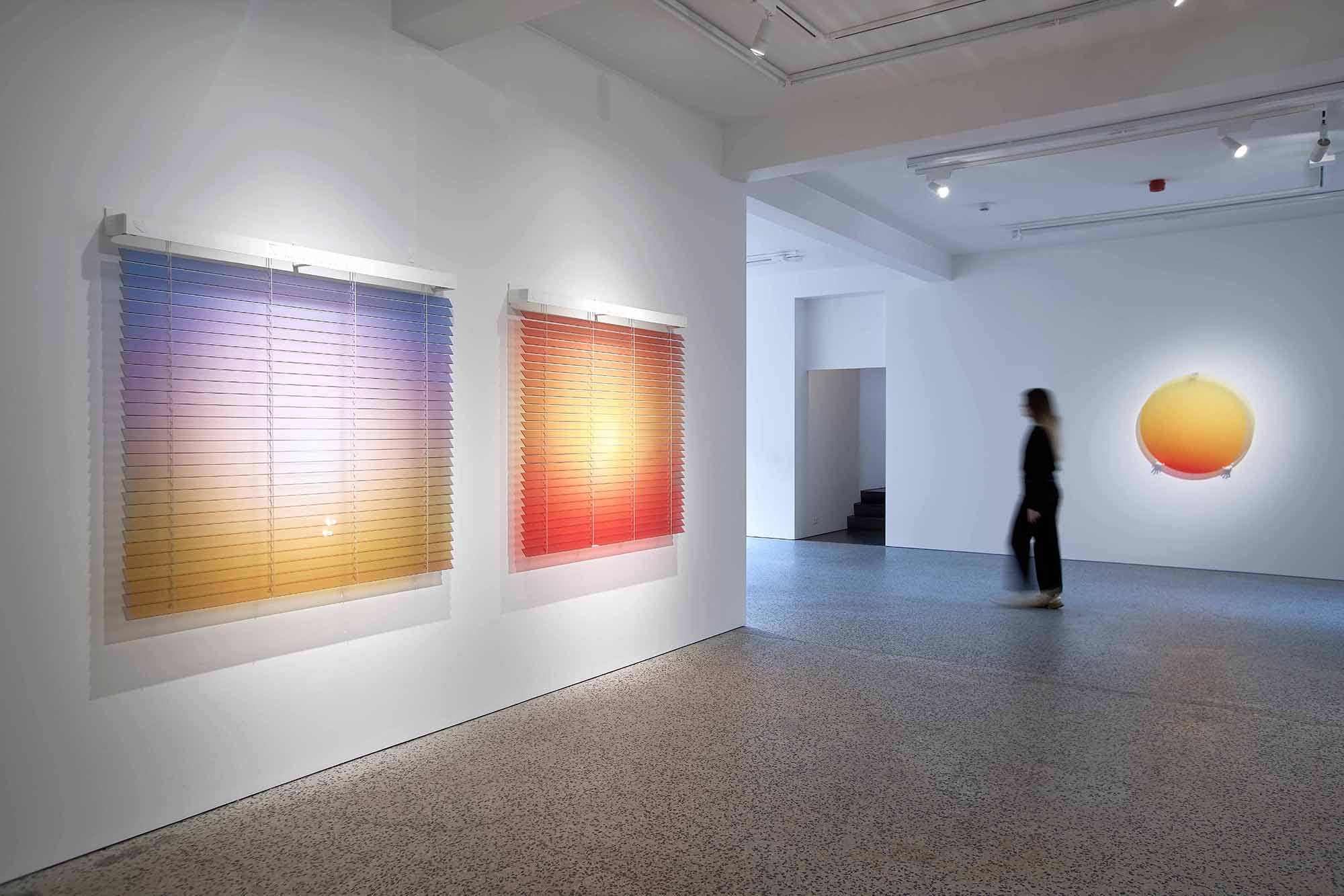 Thordis Erla Zoega, Domestic Sci-fi, installation view: Everyday — blue dichroic circle visible through hanging white venetian blind panel at left — and two Forecast gradient blind works on right wall in warm amber to orange-red, BERG Contemporary, Reykjavík, 2025.
