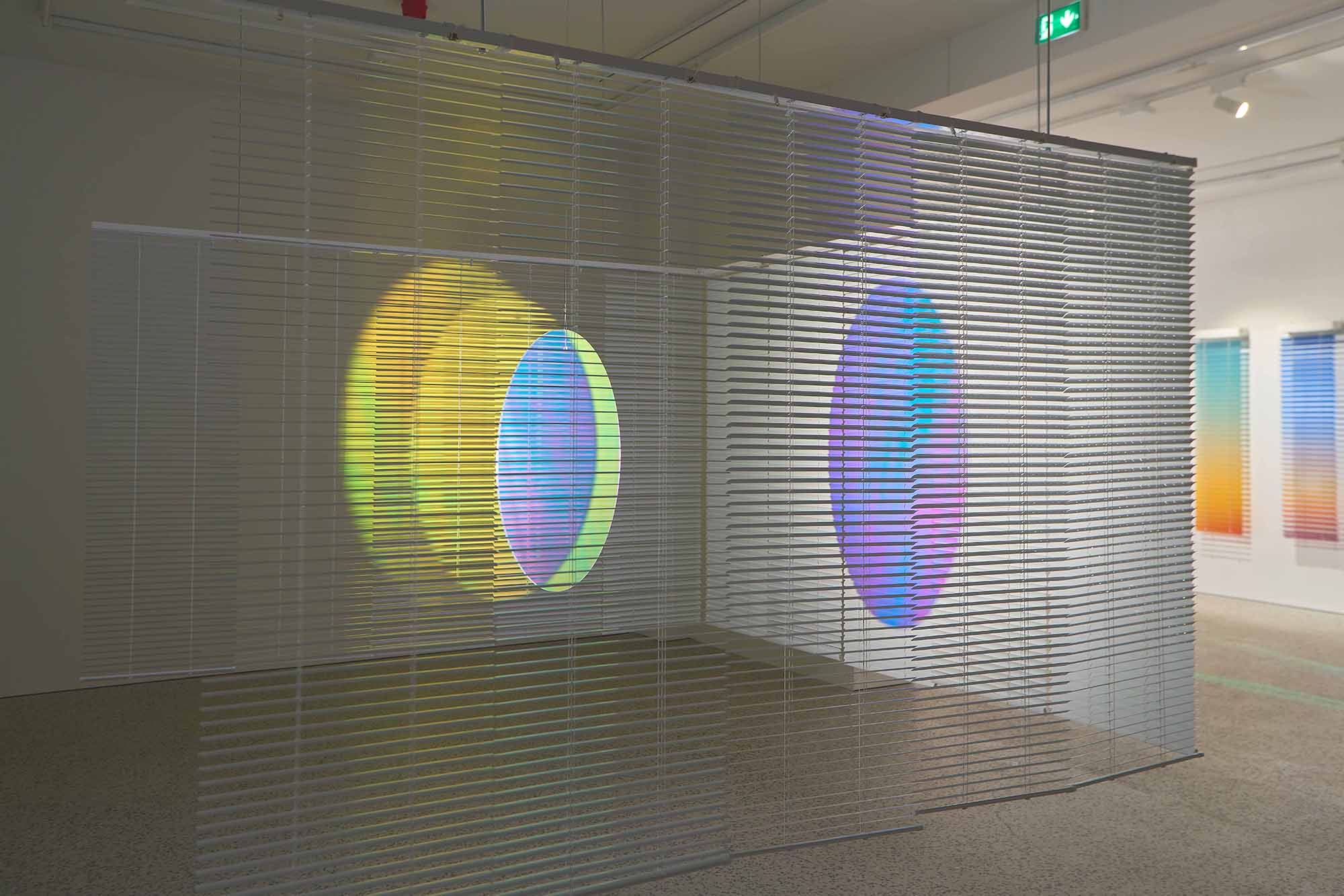 Thordis Erla Zoega, Everyday, installation view: large hanging dichroic blind panel displaying full-spectrum iridescent effect — teal, yellow, pink, white — with rainbow-coloured light rays cast across the gallery floor below, BERG Contemporary, Reykjavík, 2025.