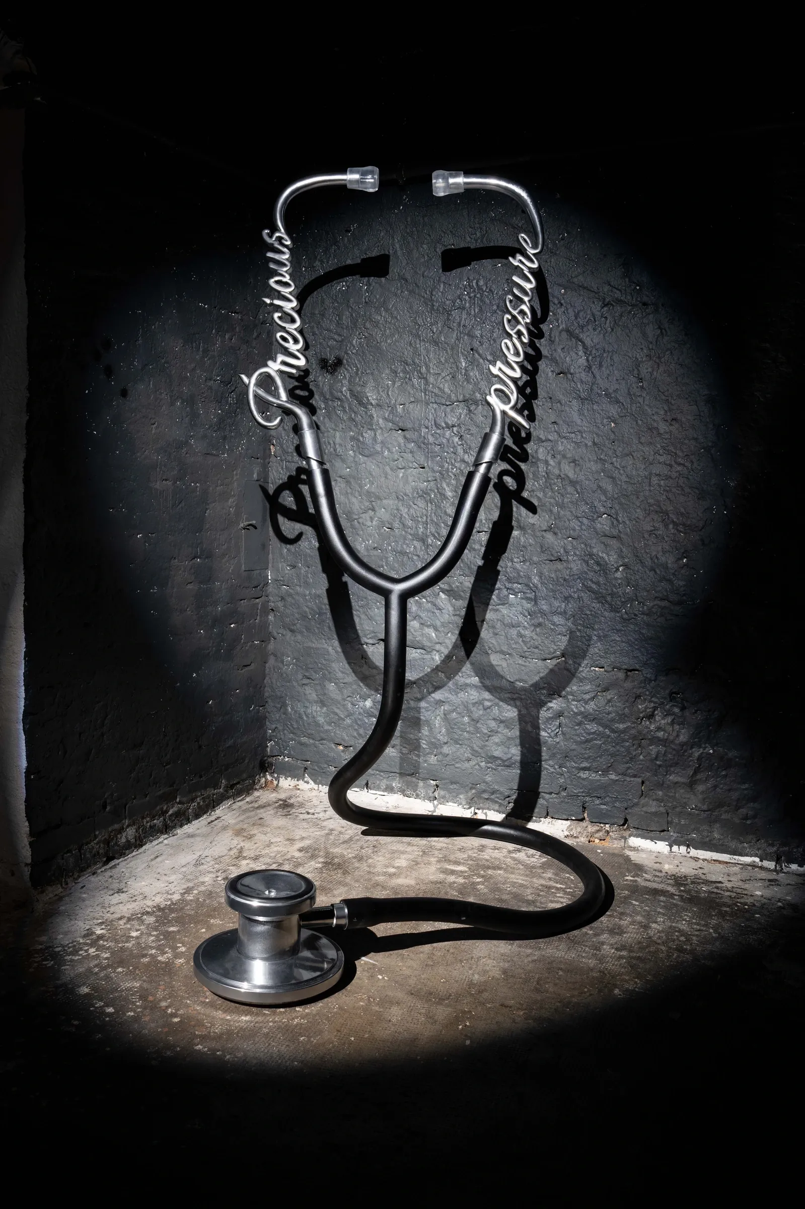 Joschua Yesni Arnaut, Precious Pressure, Galerie 3AP, Frankfurt, 2026, oversized stethoscope sculpture in spotlight against dark wall with script lettering