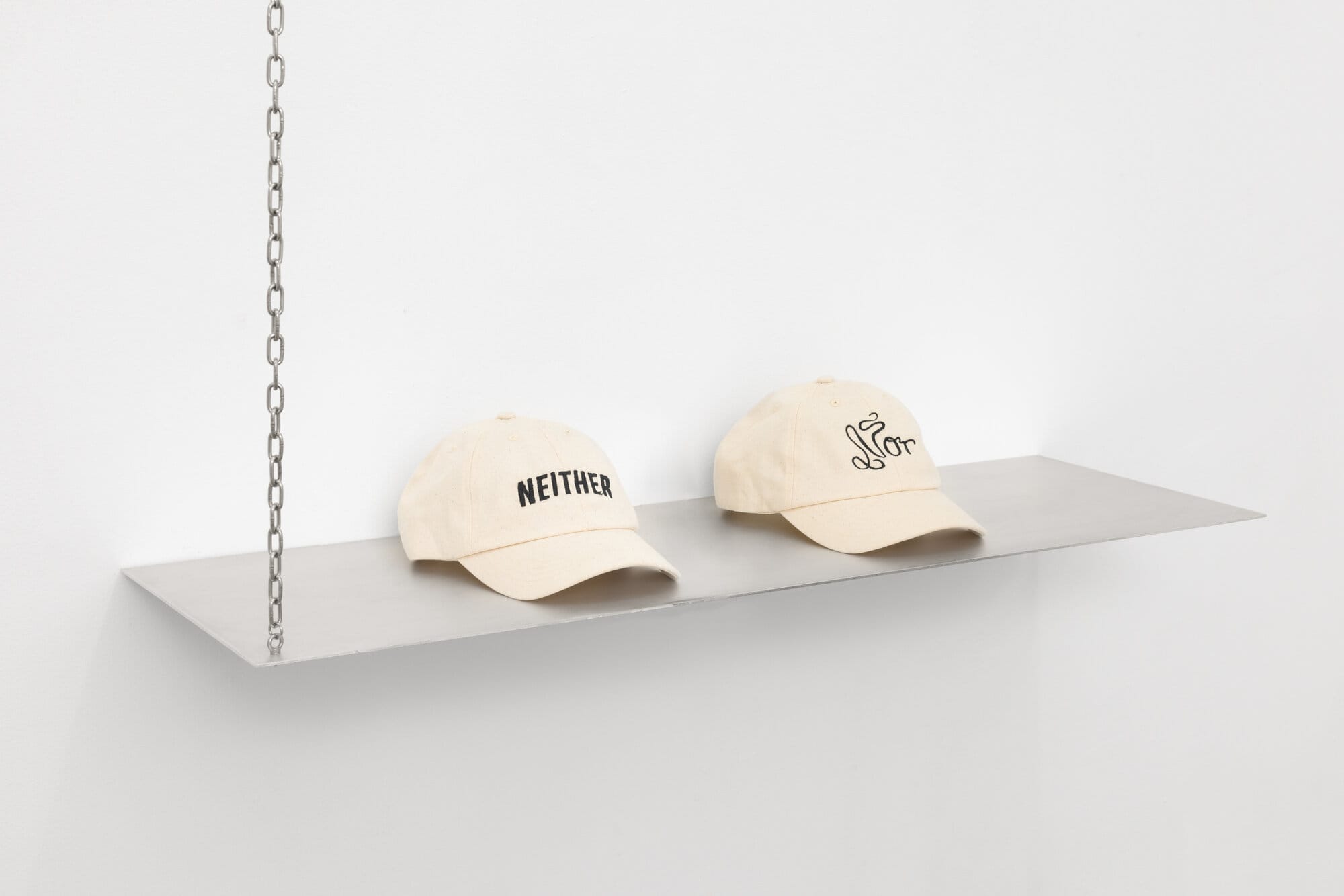 lom-of-LaMa, two cream-colored baseball caps on suspended metal shelf, one embroidered with "NEITHER" in block letters, one with "Nor" in cursive script, hanging from silver chain, Call Me We, SUPERRAUM Dortmund, 2026